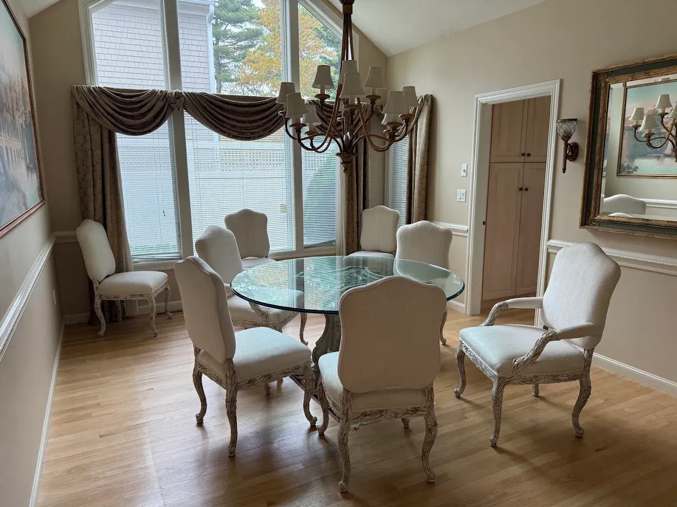 Elegant dining room with a glass table, white chairs, chandelier, and large window with drapes.