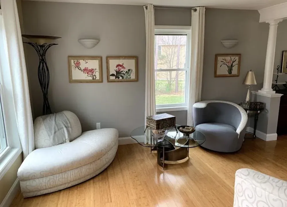 Modern living room with two gray chairs, a white chaise lounge, glass table, and framed wall art.