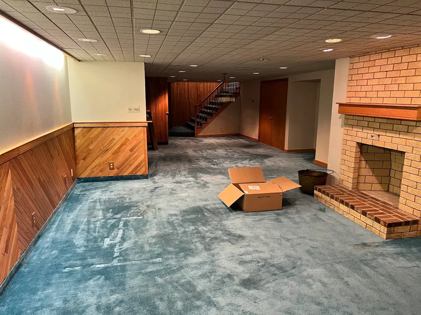 Empty basement room with blue carpet, wood paneling, brick fireplace, and cardboard box on the floor