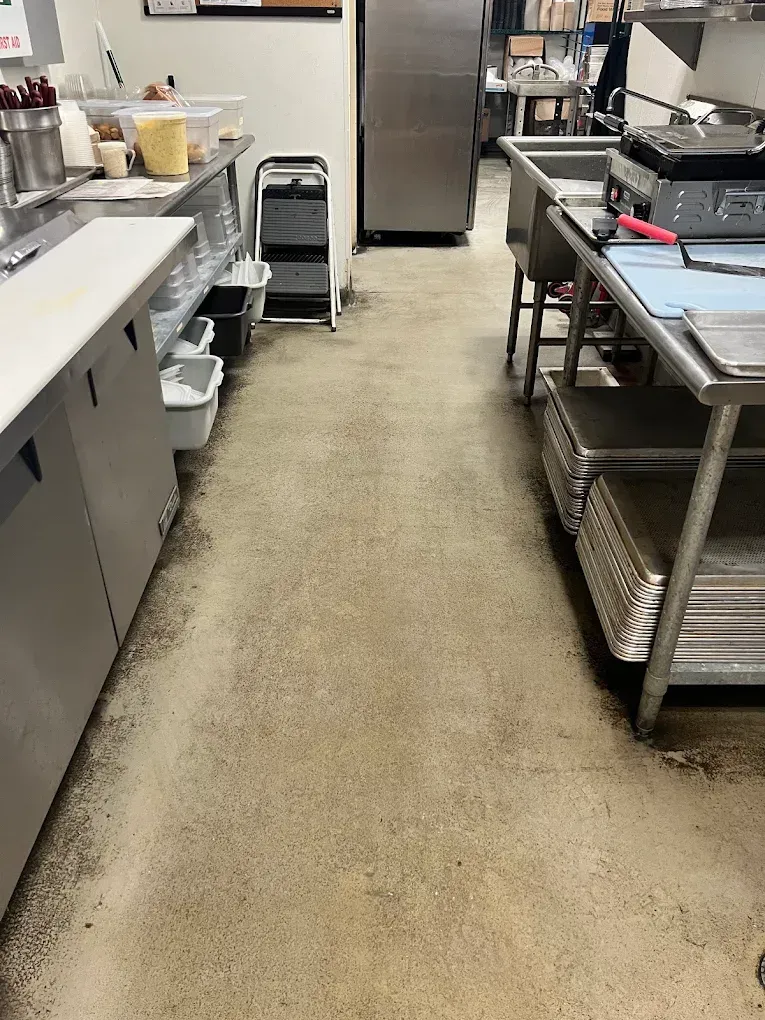 Commercial kitchen aisle with stainless steel counters, equipment, and a refrigerator at the back.