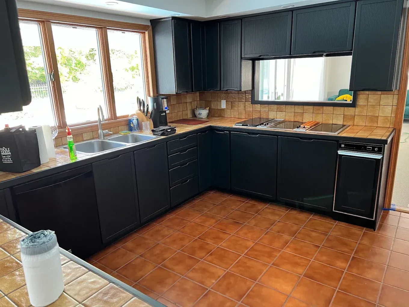 Black L-shaped kitchen with terracotta tile floor, large window, sink, and stainless stove.