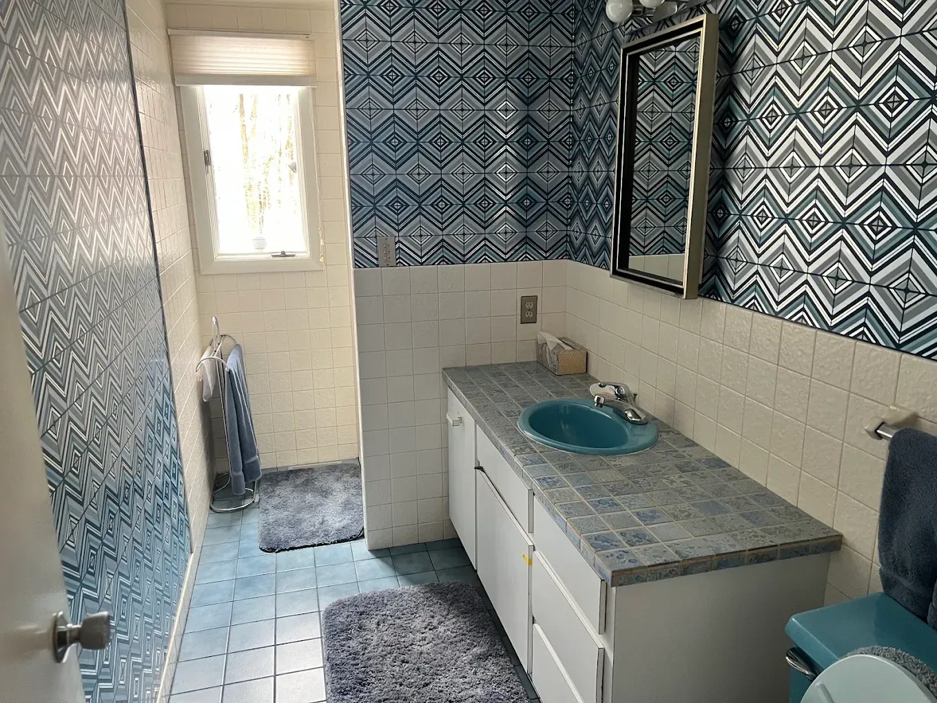 Narrow blue-and-white bathroom with patterned tile, vanity sink, mirror, and window at the far end.