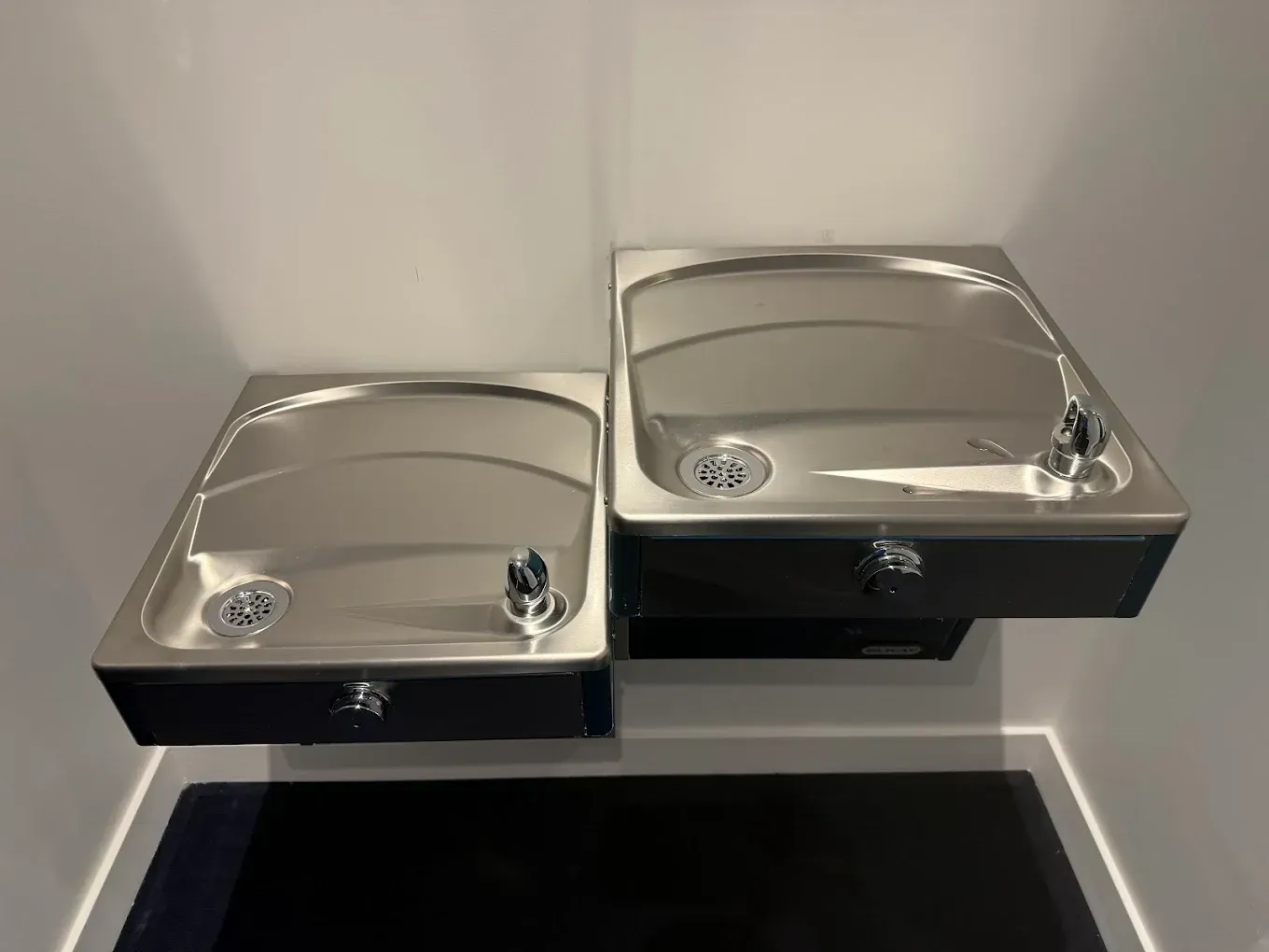 Two stainless steel drinking fountains mounted in a corner above a black floor mat