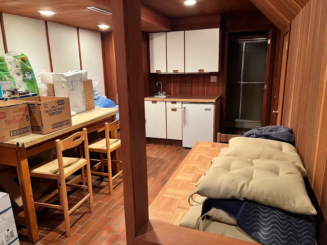 Narrow wood-paneled room with bunk bed, chairs, table, cabinets, and a folded futon mattress.