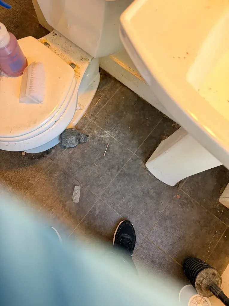 Bathroom floor with toilet, sink, and a person’s shoe visible near the bottom.
