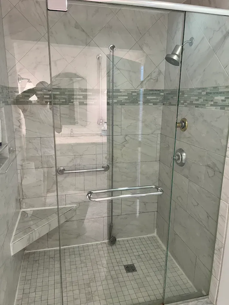 Glass walk-in shower with chrome grab bars, bench seat, handheld shower, and tiled floor.