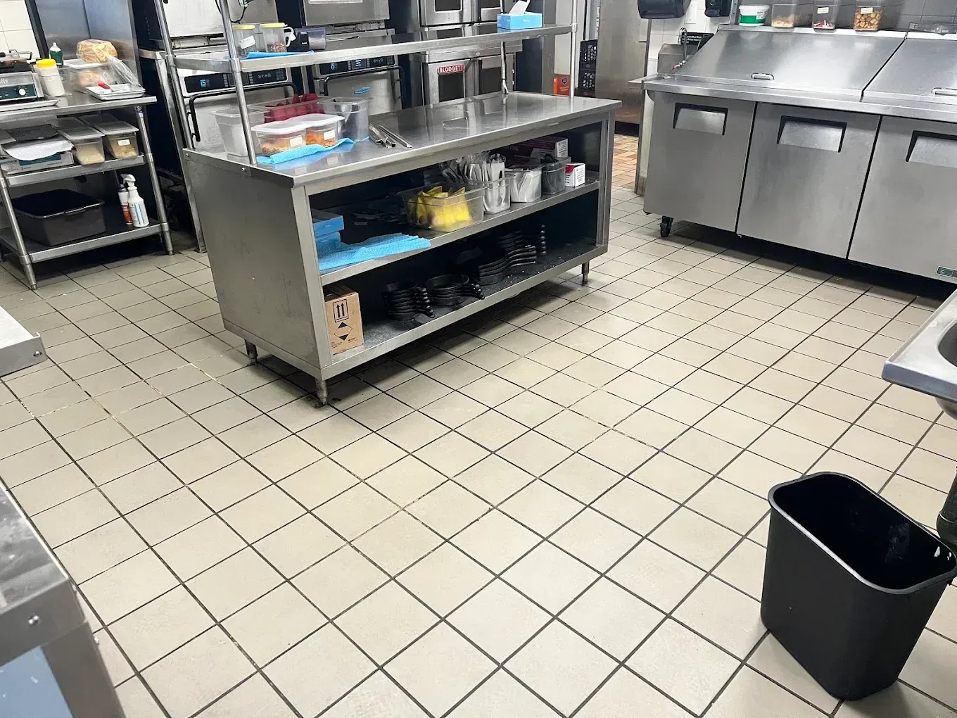Commercial kitchen with stainless steel counters, tiled floor, and a black trash bin in the foreground.