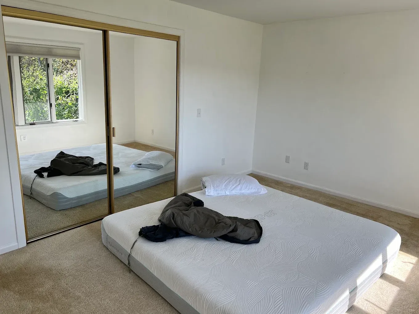 Bright bedroom with white walls, mirrored closet, and an unmade bed with clothes on it.