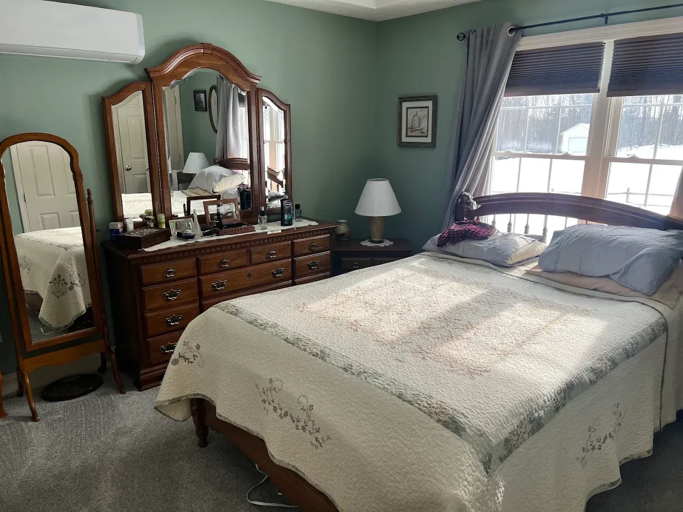 Bedroom with a made bed, wooden dresser and mirror, and a window with blinds and curtains.