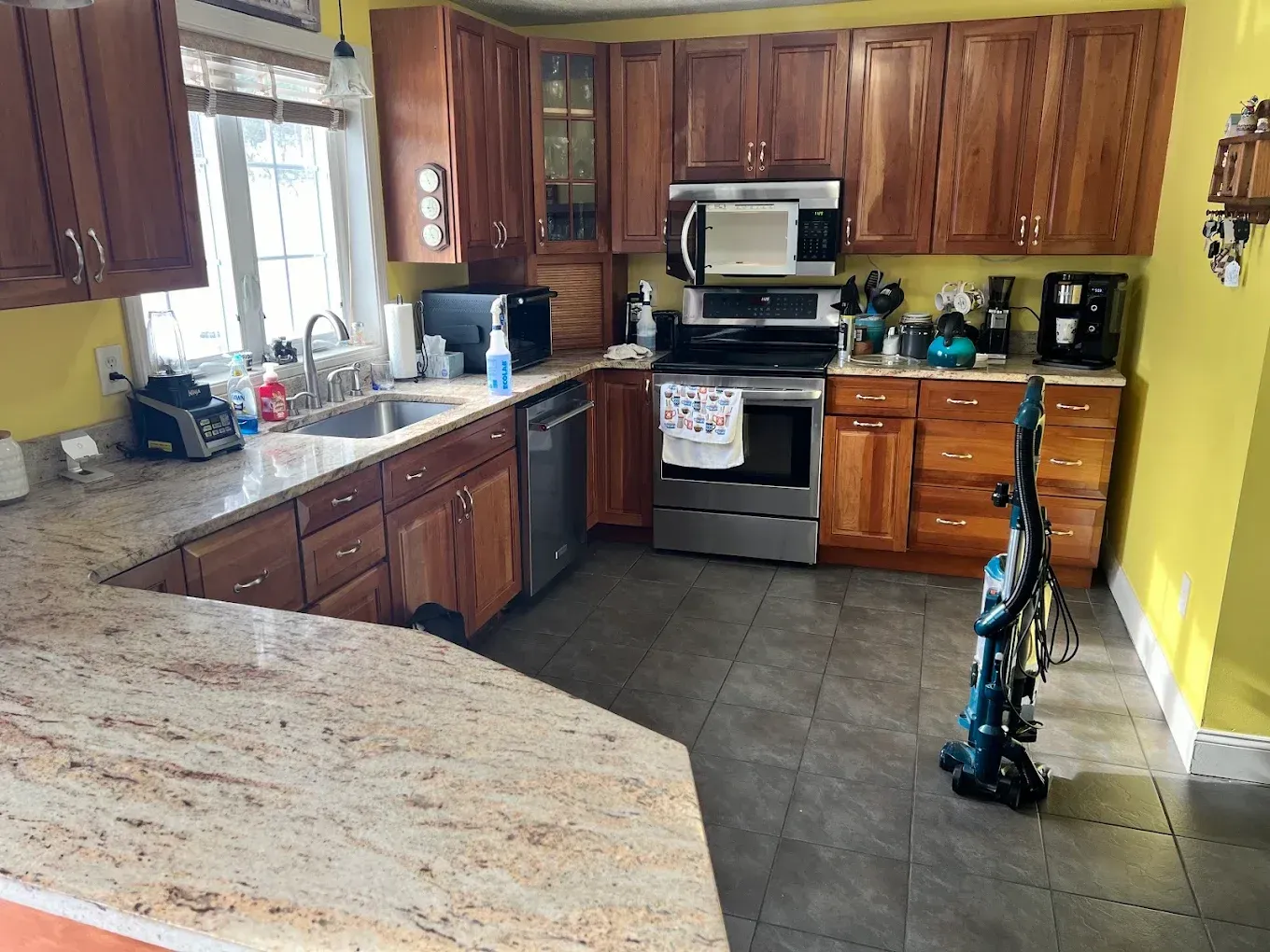 Bright kitchen with wood cabinets, granite counters, stainless stove, and a vacuum on the tile floor.