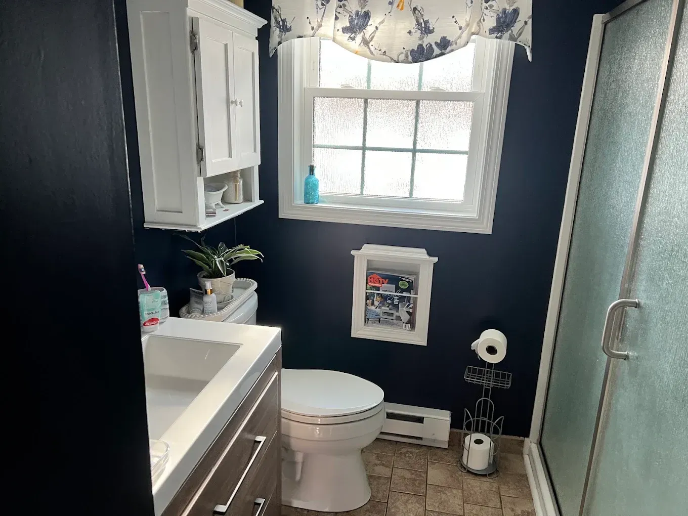 Small bathroom with a toilet, sink vanity, frosted shower door, and window with floral curtain.