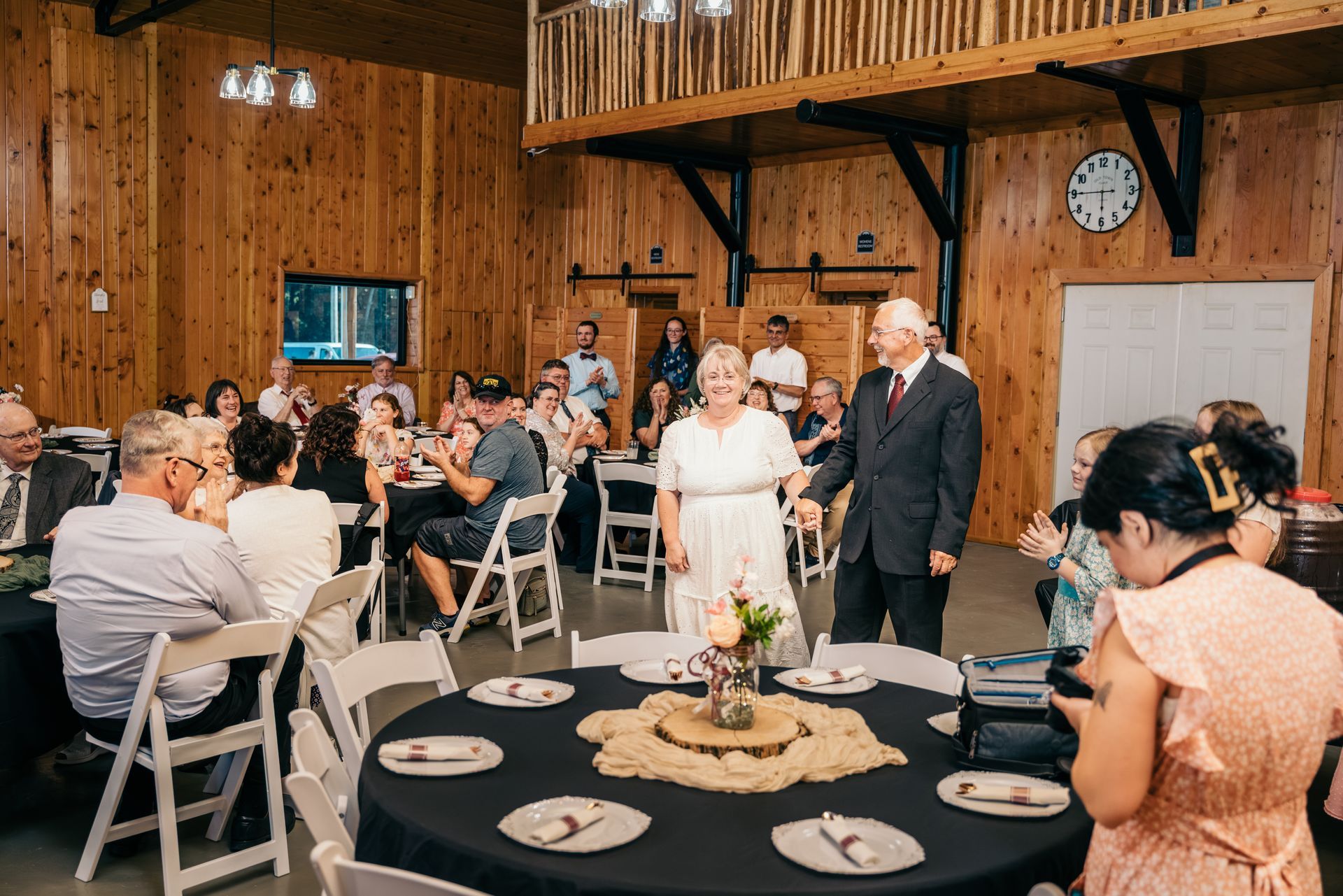 Wedding guests seated in a rustic hall as an older couple walks toward the front table