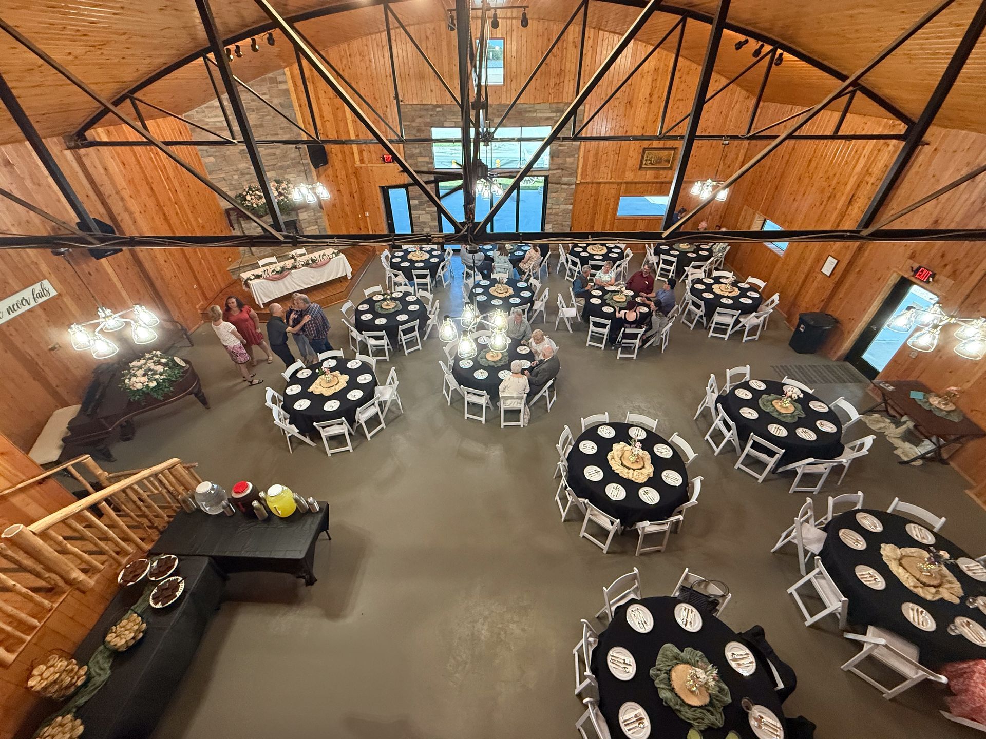 High-angle view of a rustic event hall with wooden walls, black-clothed round tables, and people gathering. High-angle view of a rustic event hall with wooden walls, black-clothed round tables, and people gathering.