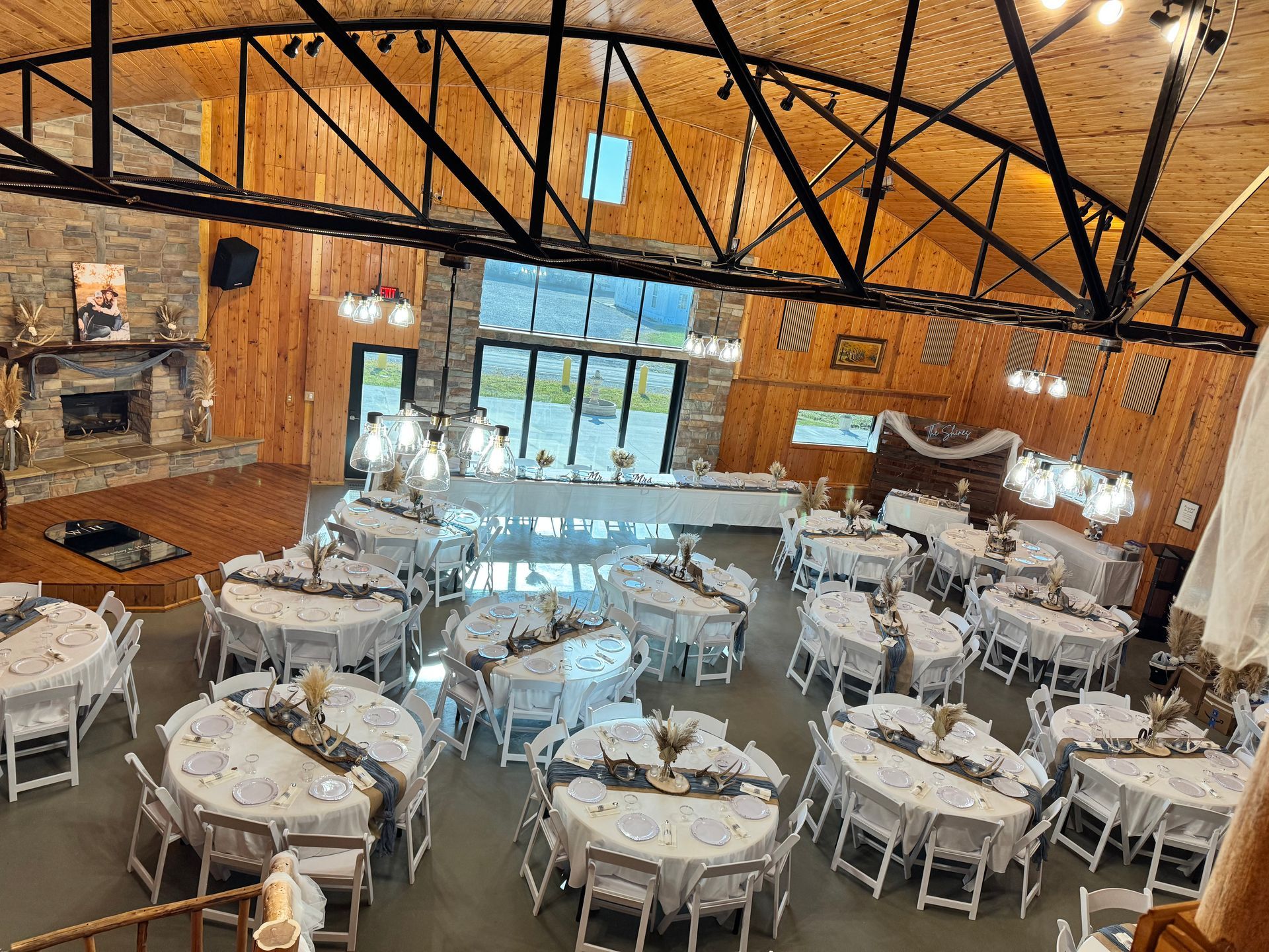 High-angle view of a rustic event hall set up with round tables for a dinner, featuring wooden walls and a stone fireplace. High-angle view of a rustic event hall set up with round tables for a dinner, featuring wooden walls and a stone fireplace.