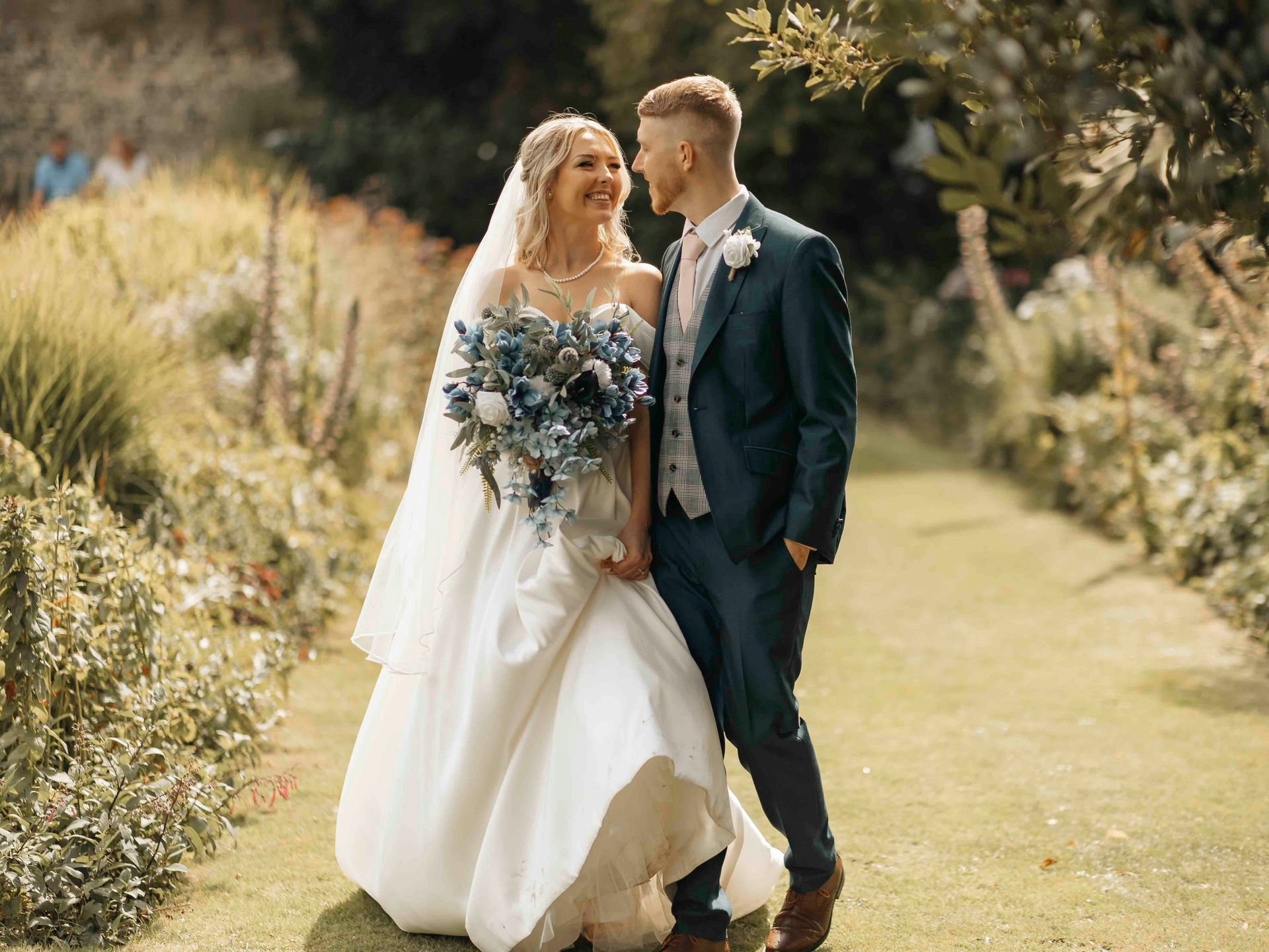 A bride and groom are walking down a path in a garden holding hands.