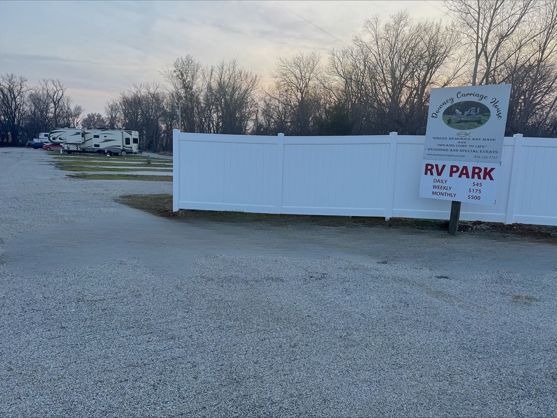 Gravel RV park with white fence and sign; RVs parked in the distance, trees and overcast sky.