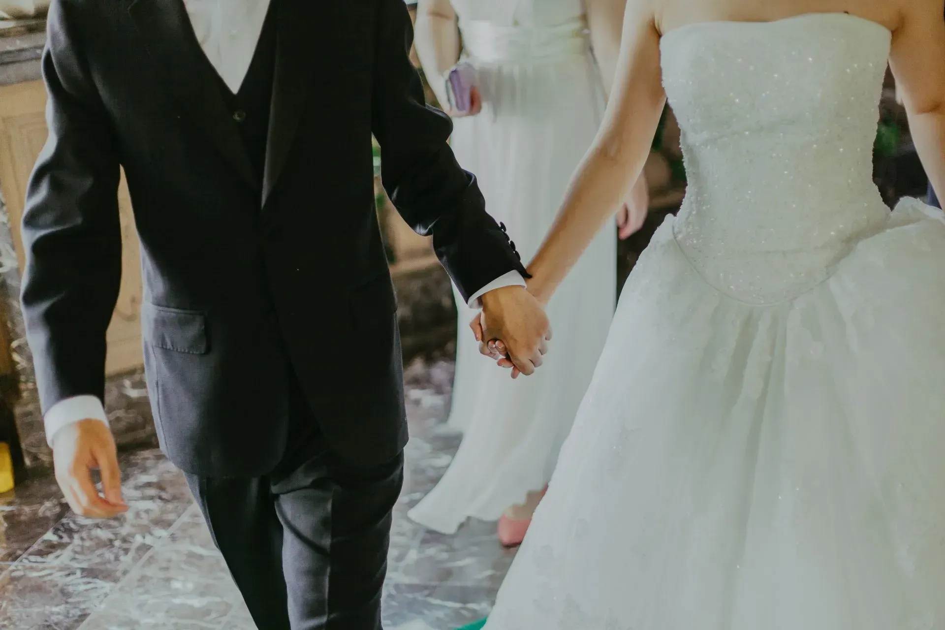 A person in a black suit and a person in a white wedding gown hold hands while walking on a marble floor.