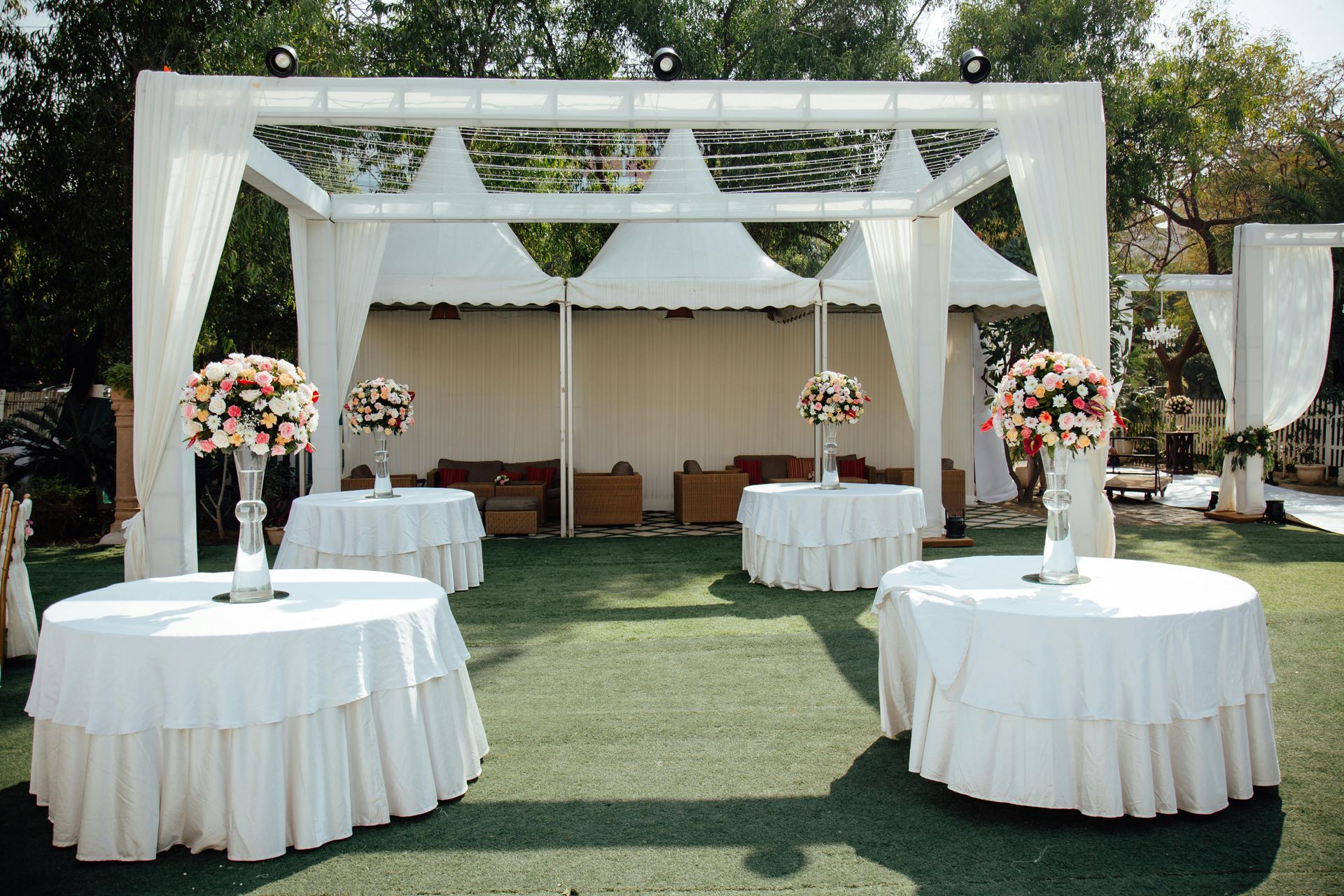 Outdoor event setup with four white-clothed round tables, each featuring a floral centerpiece, under a draped white frame.