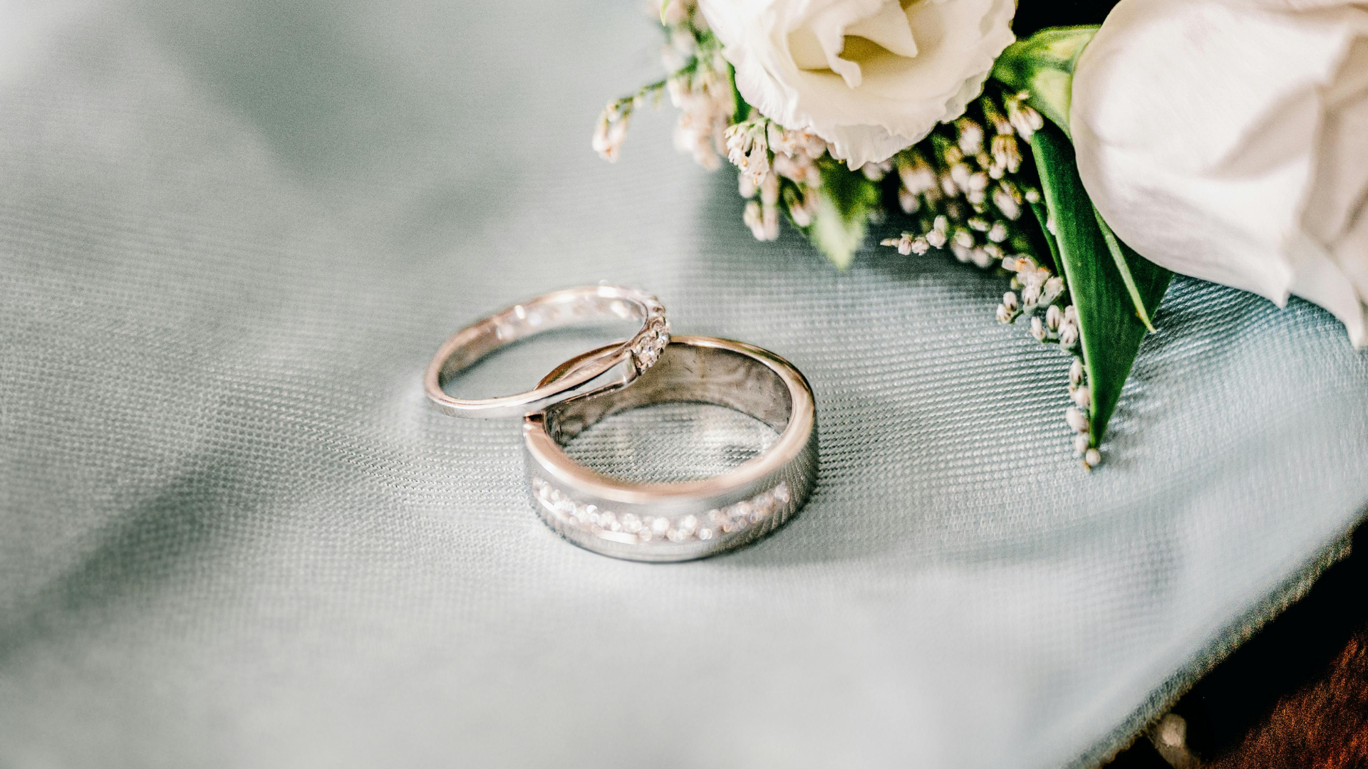 Two silver wedding rings beside white flowers on a soft gray fabric background