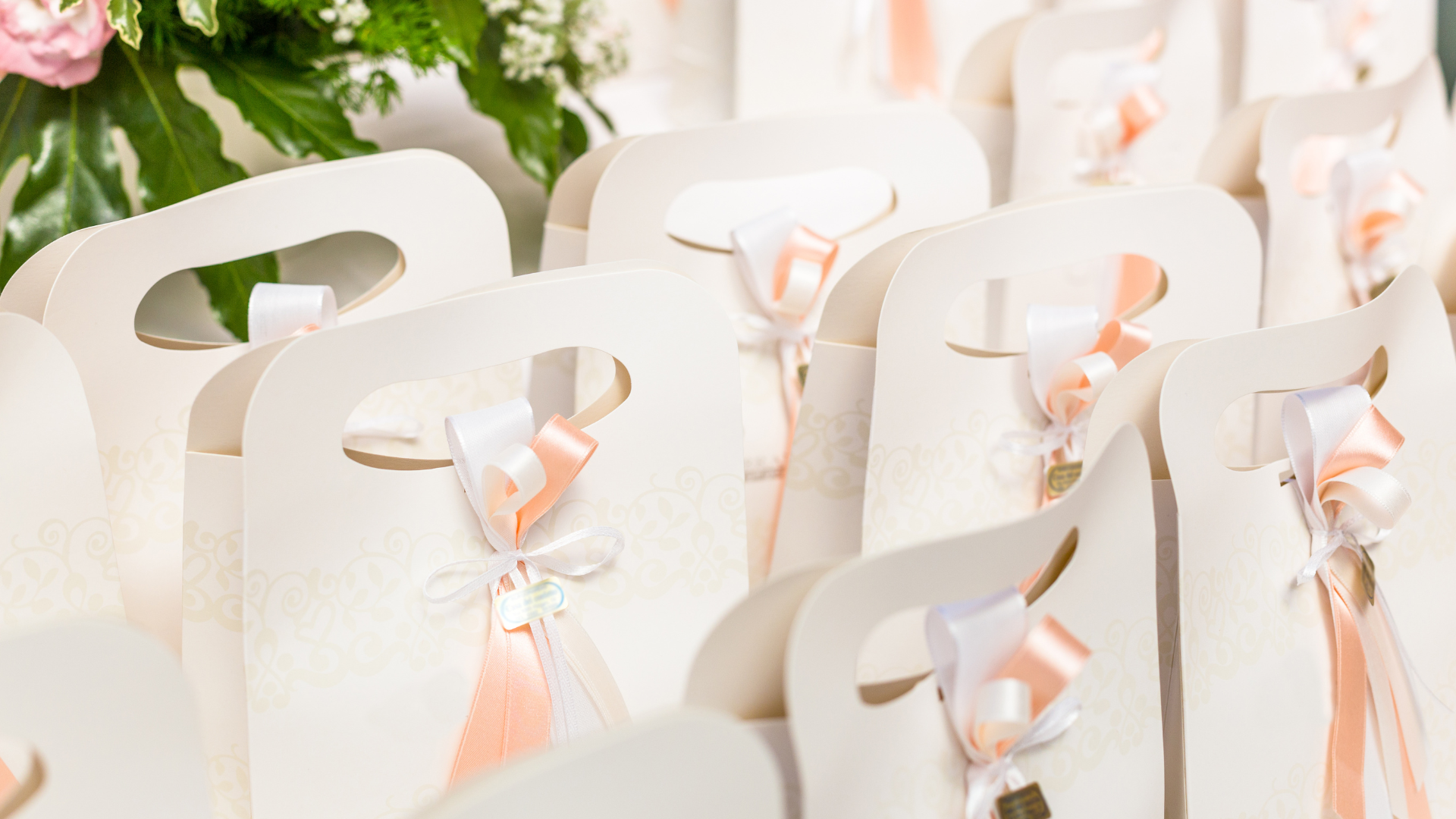 Pastel gift bags with pink ribbon bows arranged on a table, with white flowers in the background