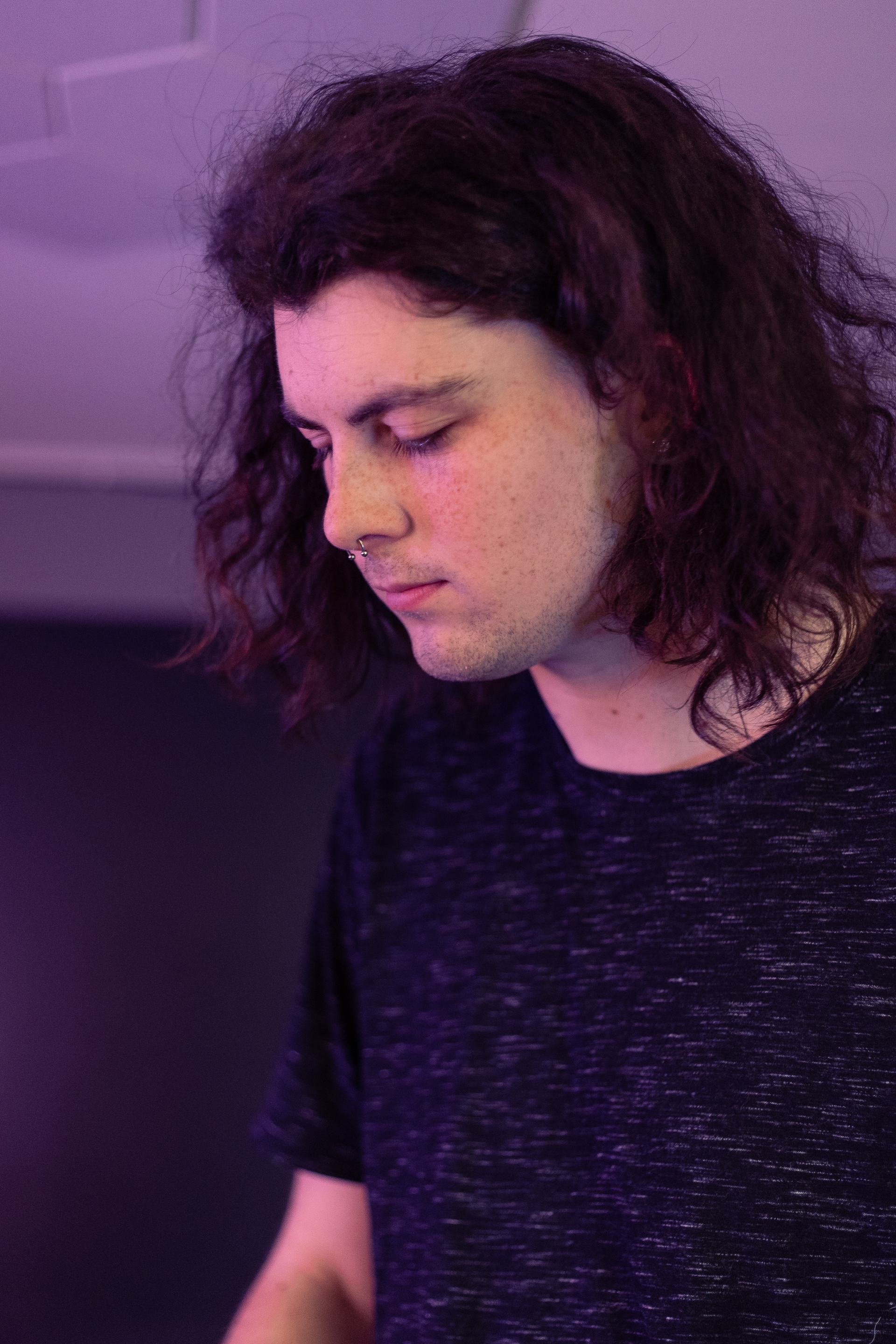 A young man with long hair is wearing a black shirt and looking down.