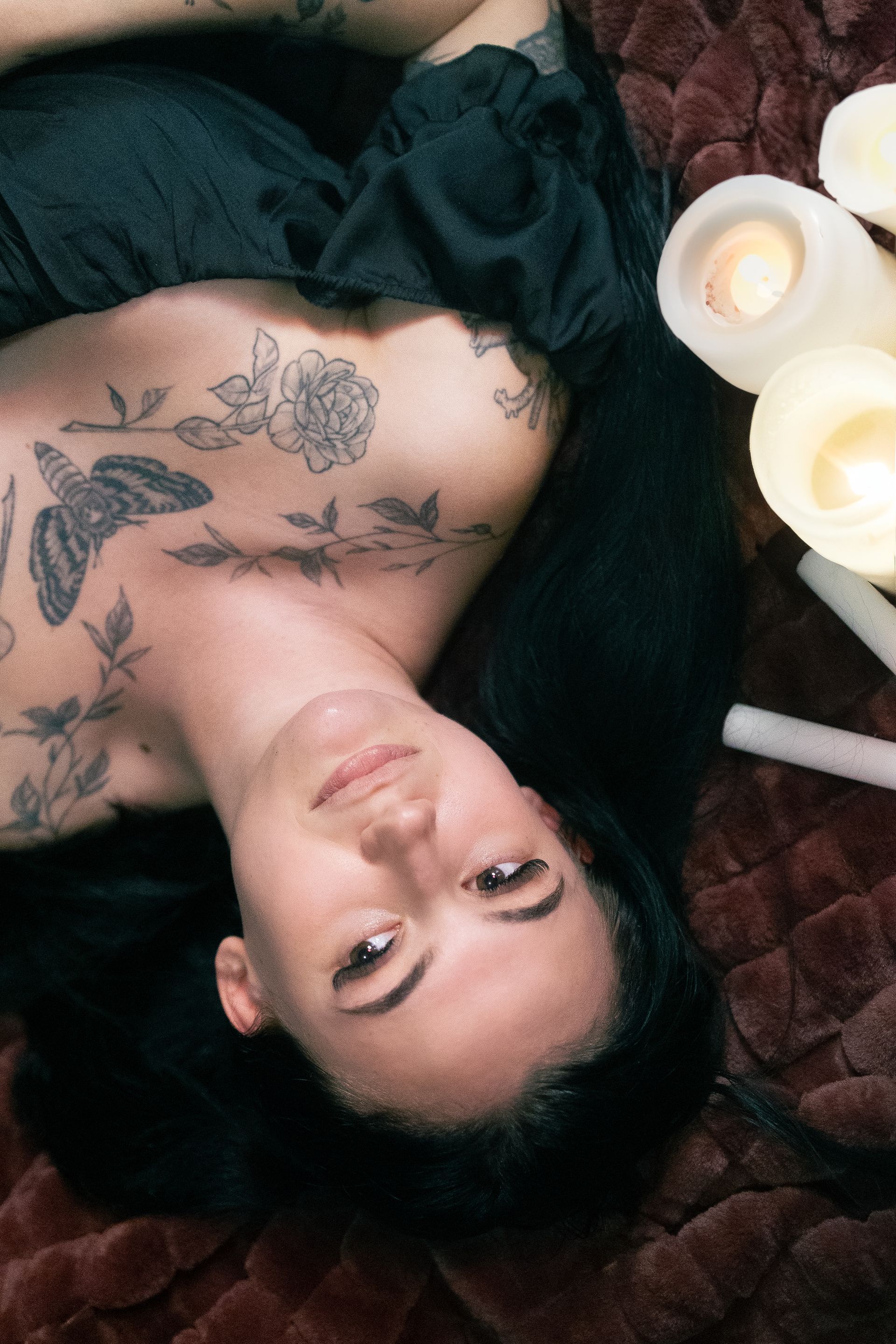 A woman with tattoos on her chest is laying on her back next to candles.