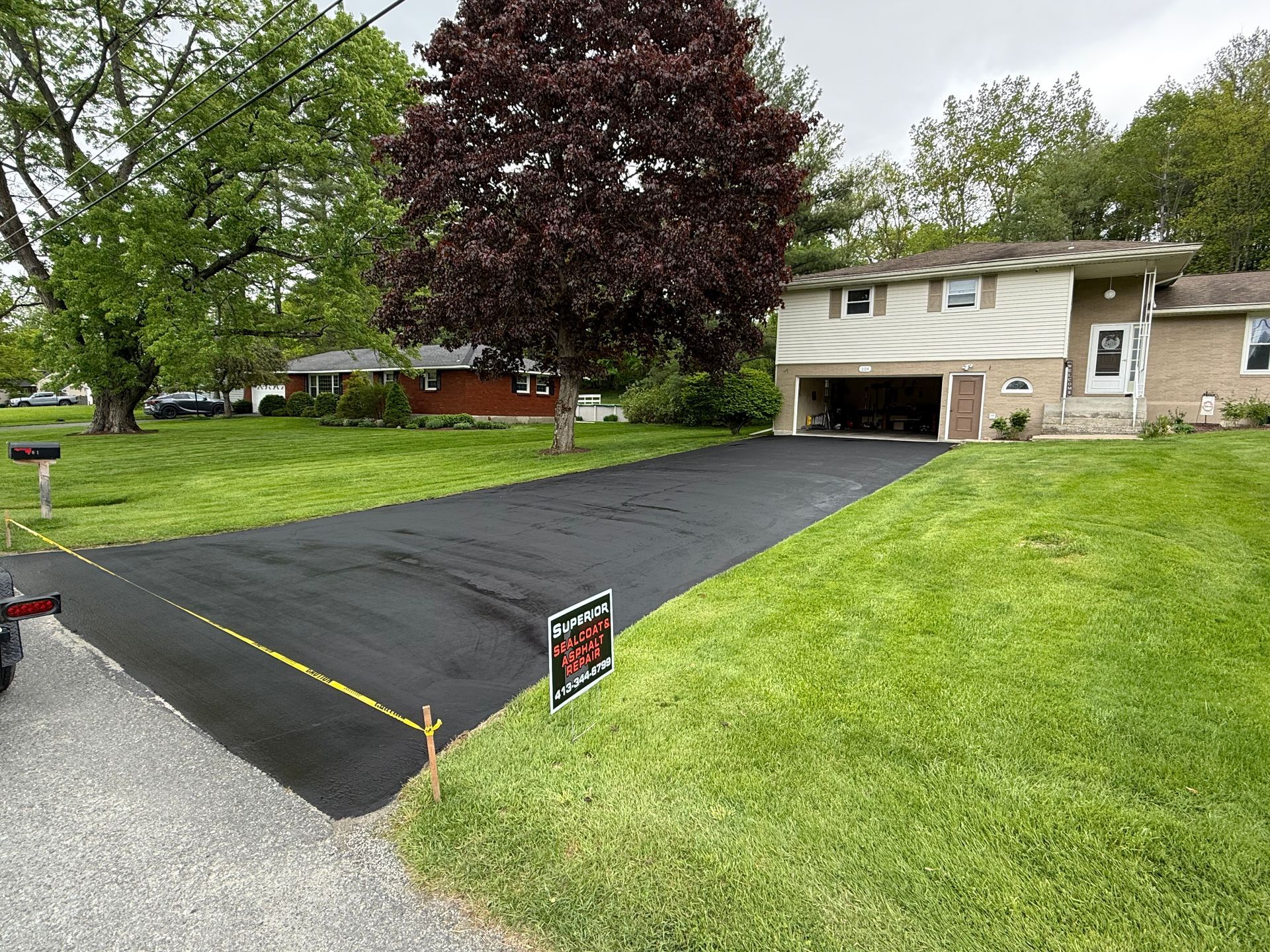 A newly paved asphalt driveway leads to a suburban split-level house on a grassy lot with mature trees.