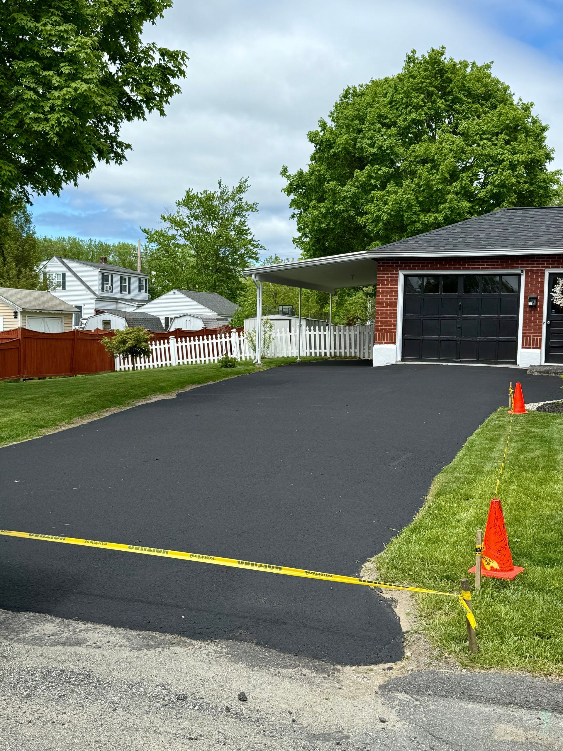 A freshly paved black asphalt driveway leading to a garage, marked with a yellow caution tape and orange traffic cones.