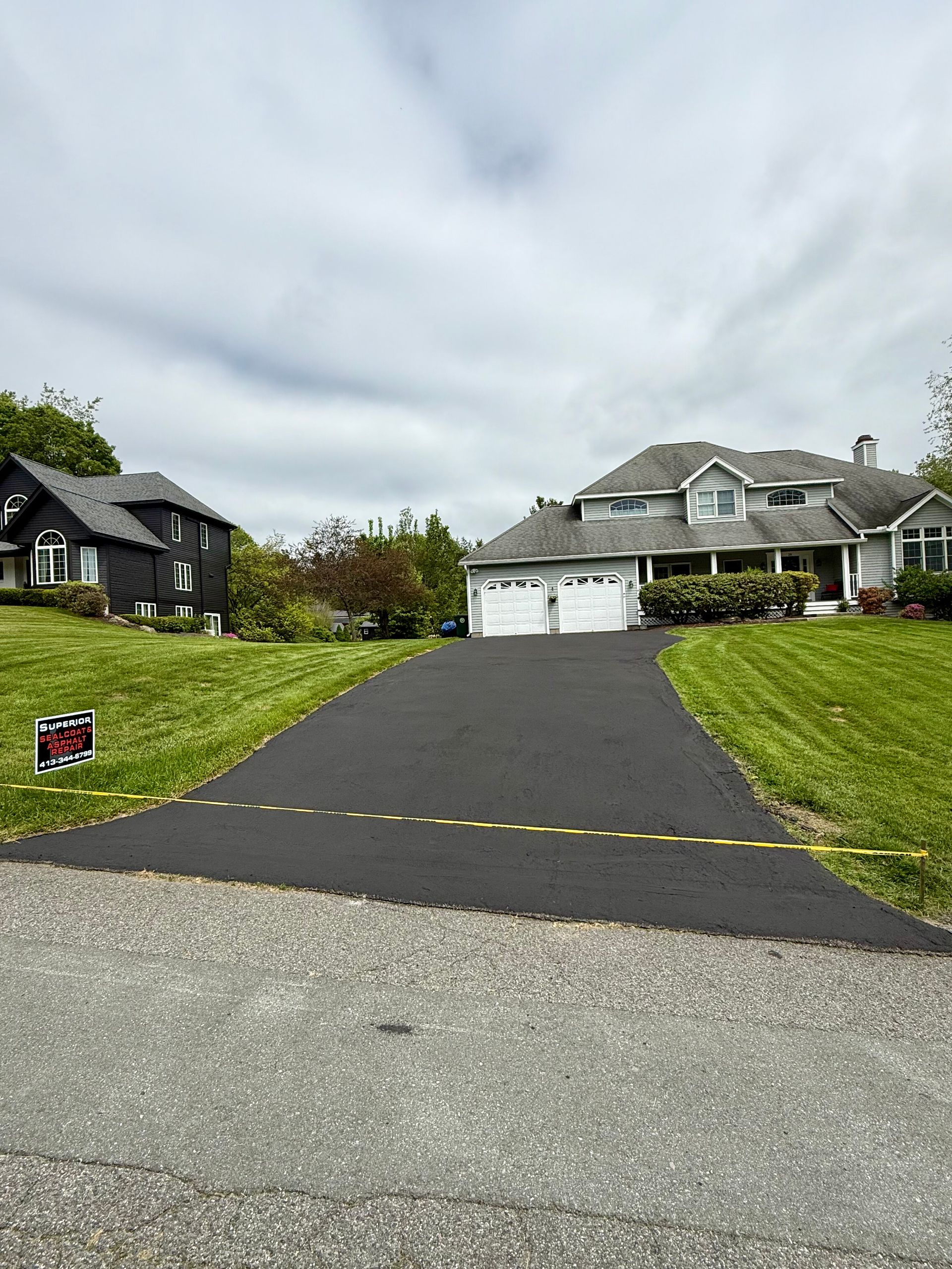 A freshly paved black asphalt driveway leads to a gray suburban house under a cloudy sky, with caution tape across it.