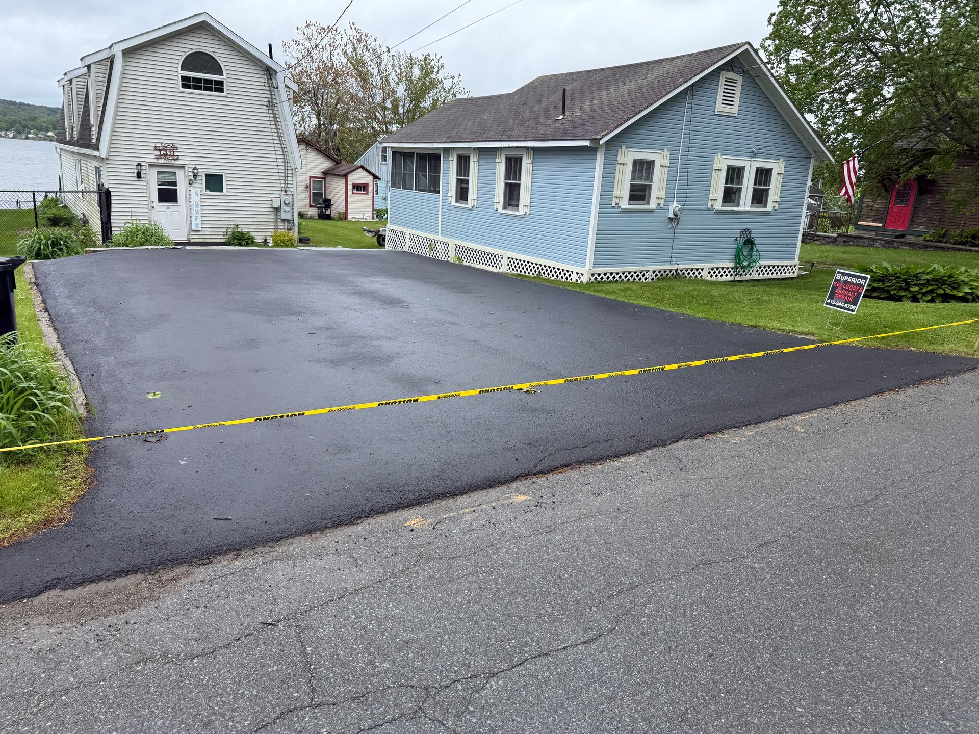 Newly paved black driveway in front of a blue house and a white barn-style building, marked off with yellow caution tape.