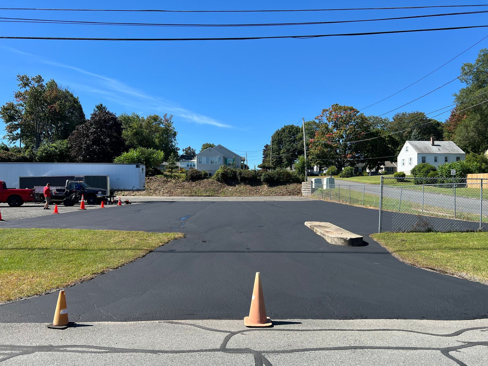 A newly paved asphalt driveway in front of a white house with orange traffic cones placed on the surface.