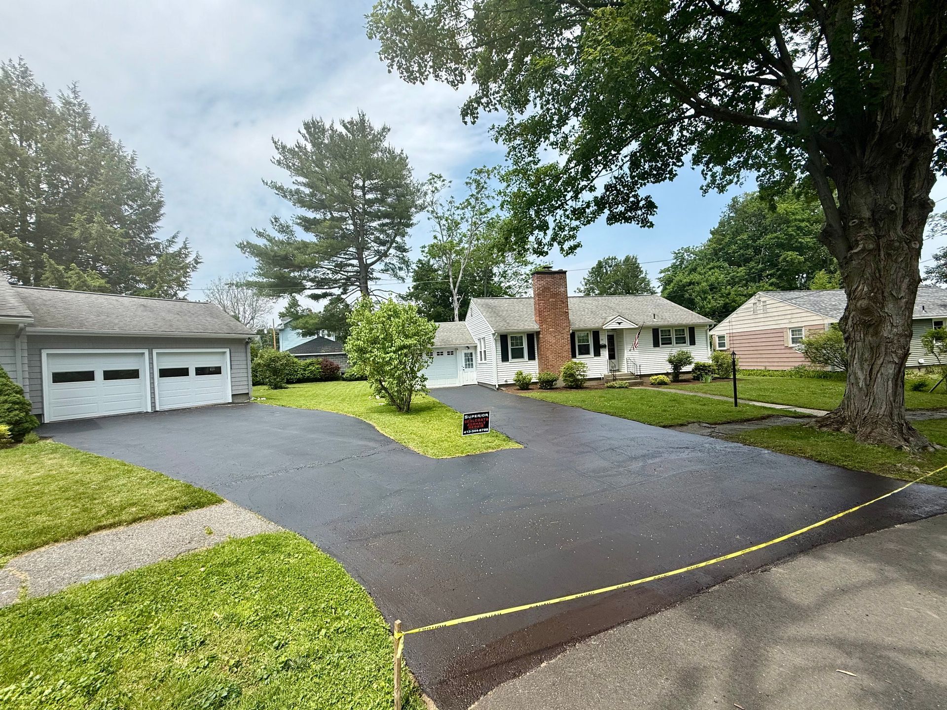 A newly paved, dark asphalt driveway leads to a white house and detached garage, separated by caution tape.