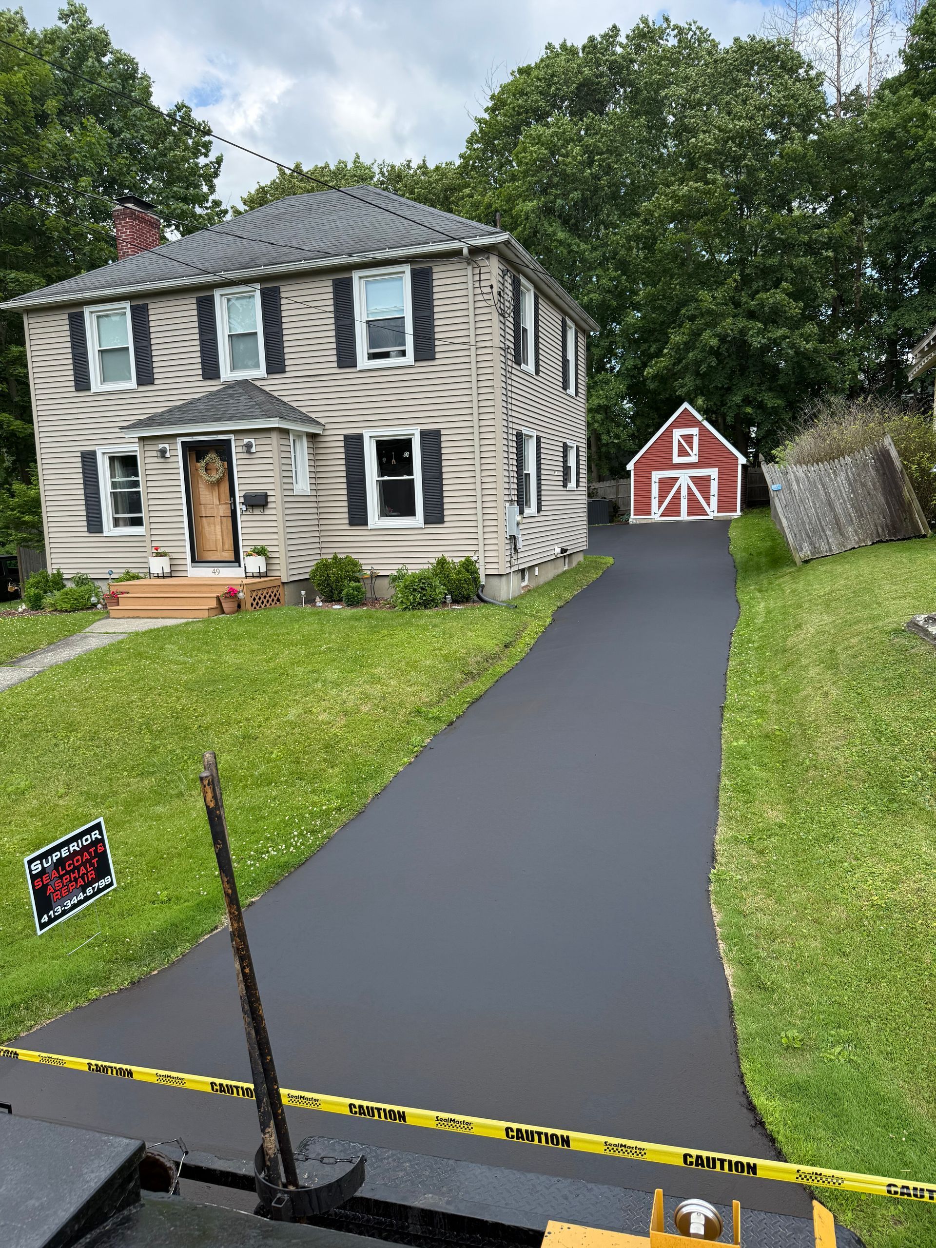 A newly paved asphalt driveway leads to a two-story beige house and a red barn-style garage on a bright, sunny day.