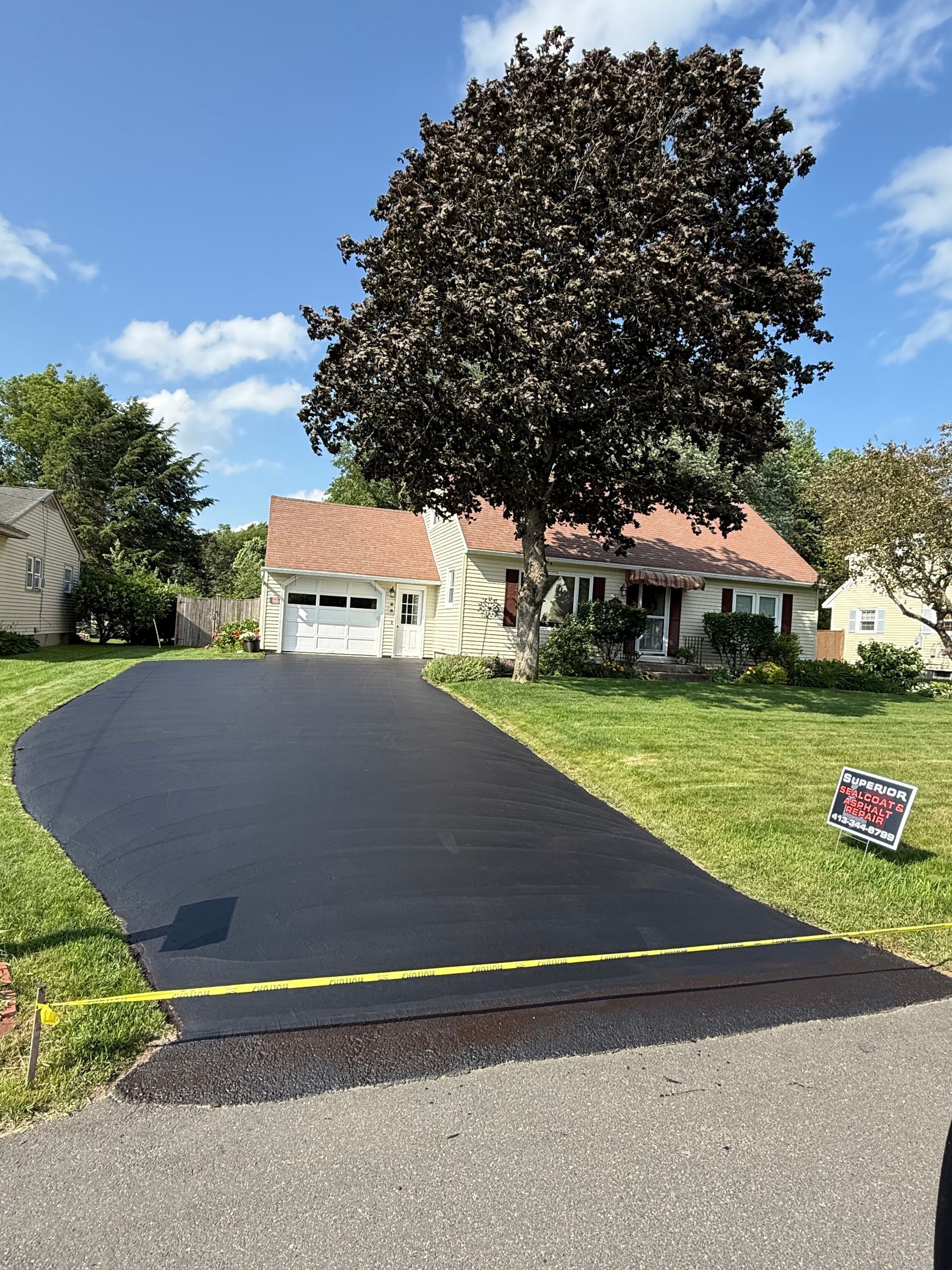 A freshly paved black asphalt driveway leads to a suburban home with a large tree in the front yard on a sunny day.