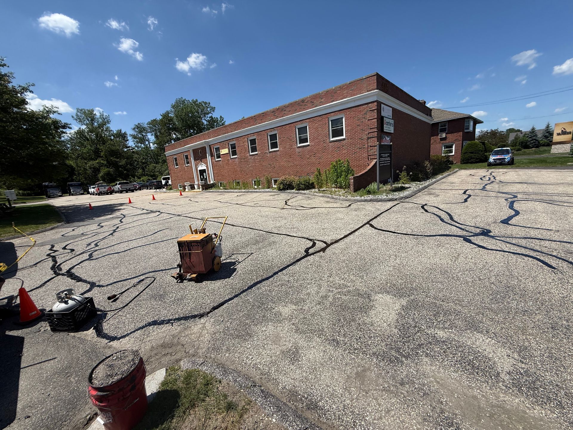 A red brick building sits behind a gravel parking lot with crack-filling sealant applied in lines across the surface.