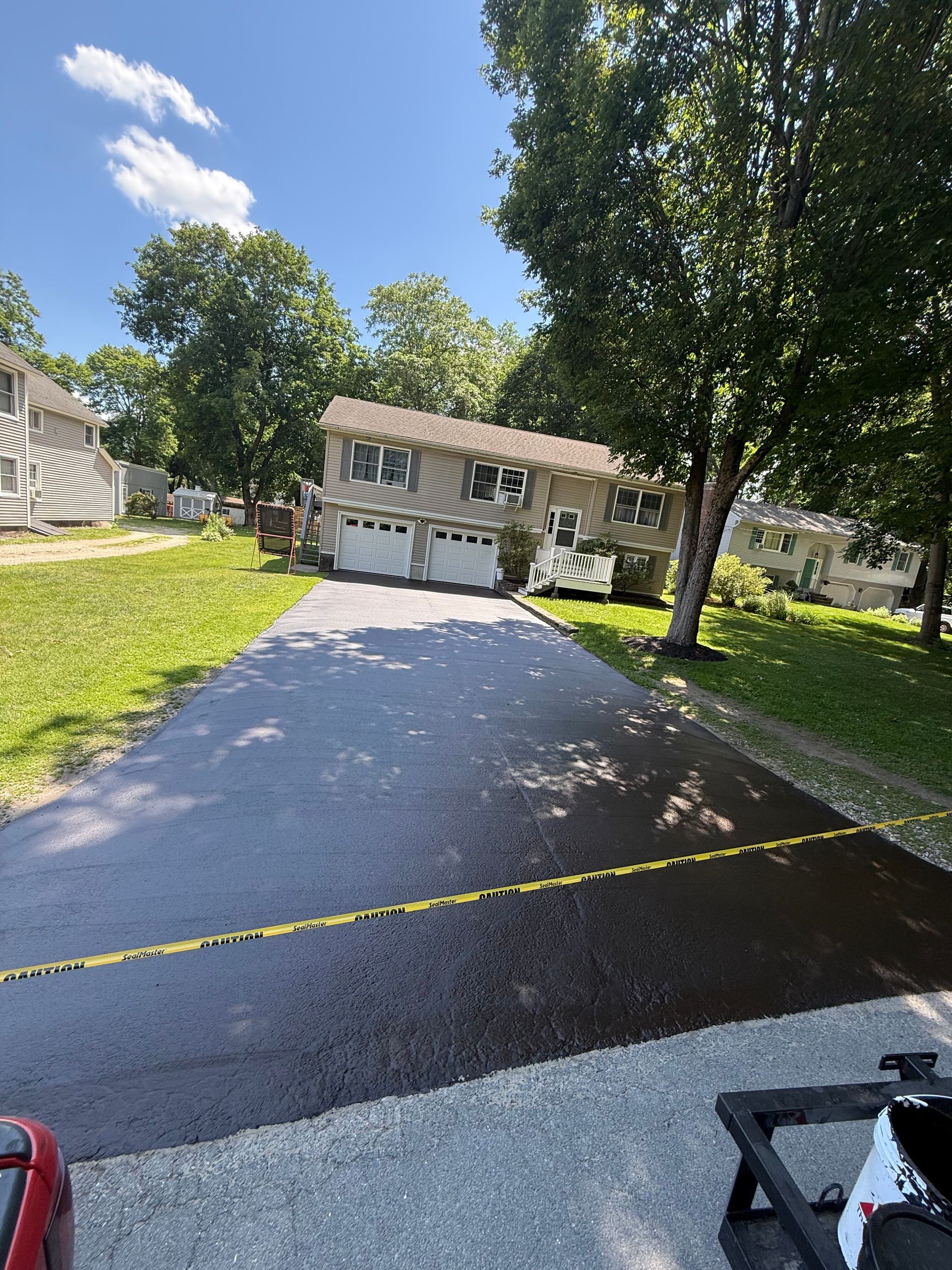 A freshly paved, dark asphalt driveway leads toward a two-story suburban house with a two-car garage on a sunny day.