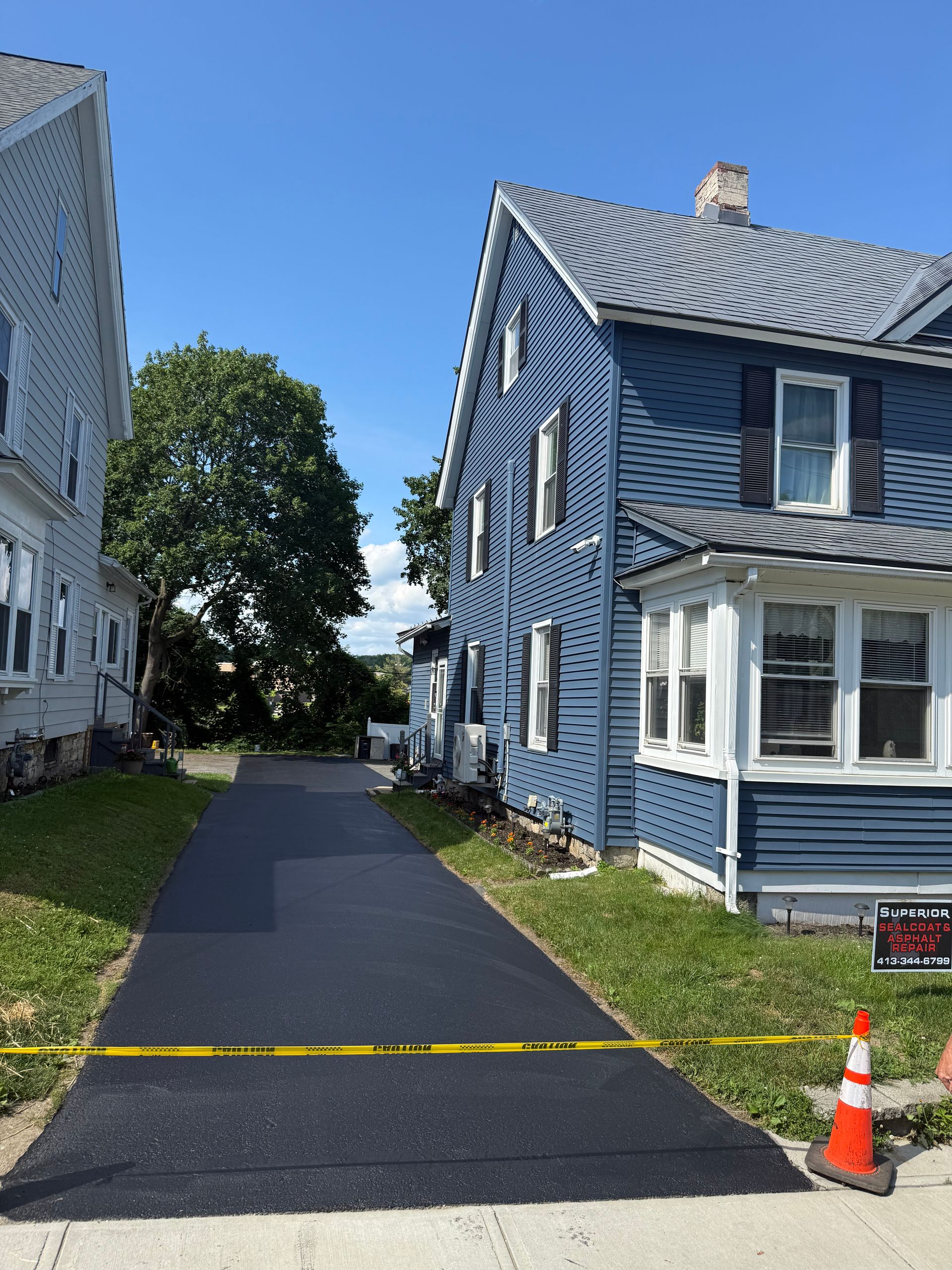 A newly paved black asphalt driveway sits between two houses, blocked off by yellow caution tape and an orange traffic cone.