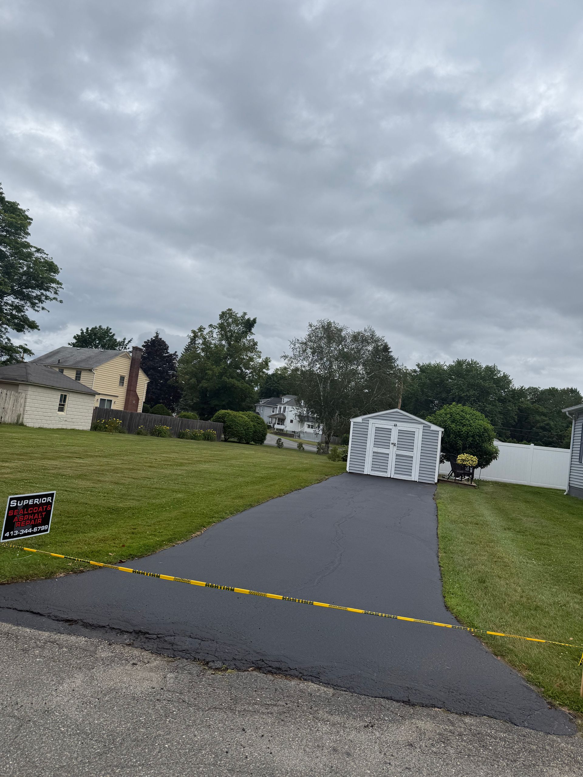A freshly paved black asphalt driveway leads to a white shed in a grassy yard under a cloudy sky.