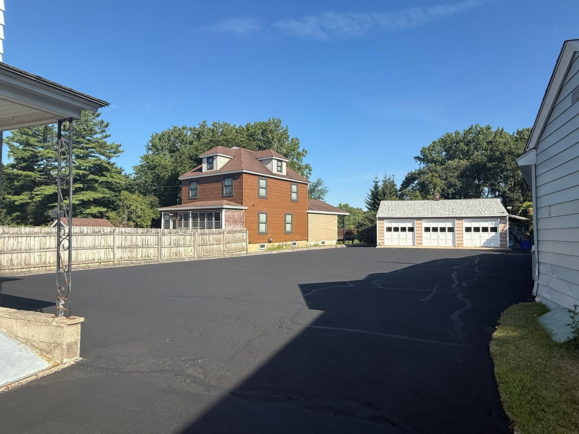 A wide asphalt driveway leads to a brown two-story house and a three-car garage under a clear blue sky.