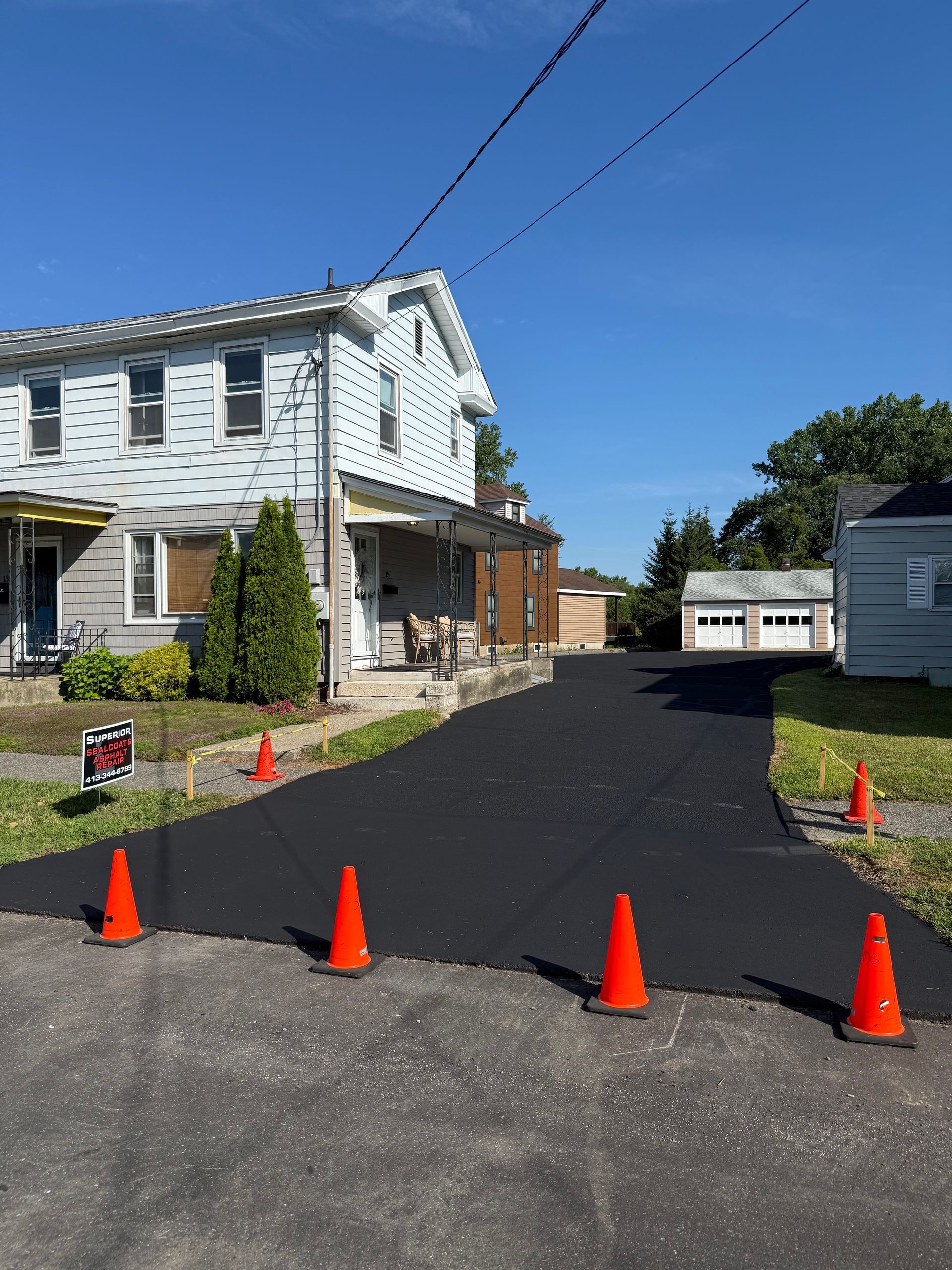 A newly paved asphalt driveway leads toward a house and a white garage, marked by orange safety cones on a sunny day.