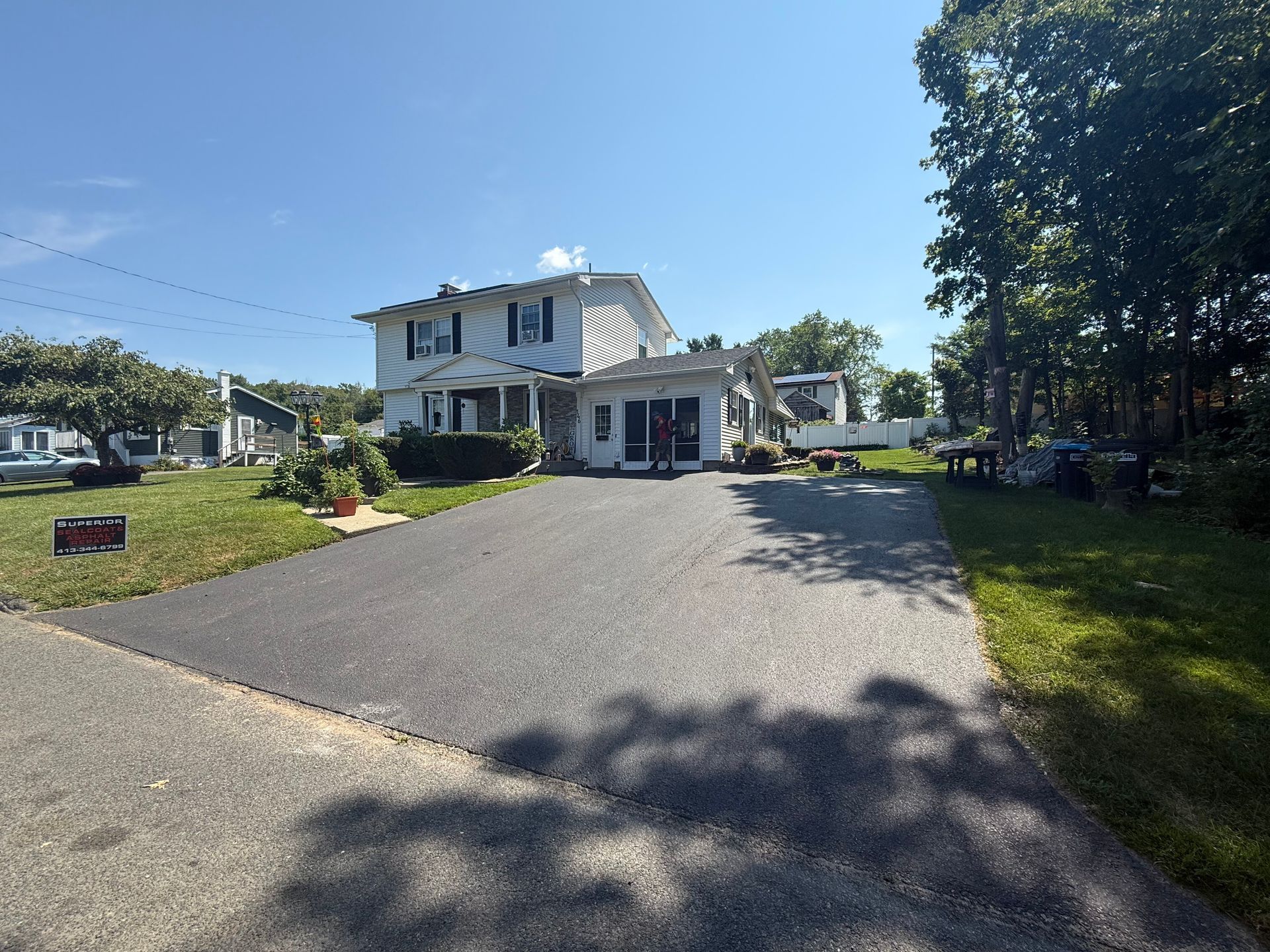 A two-story white house with black shutters sits at the end of a paved driveway under a clear blue sky.