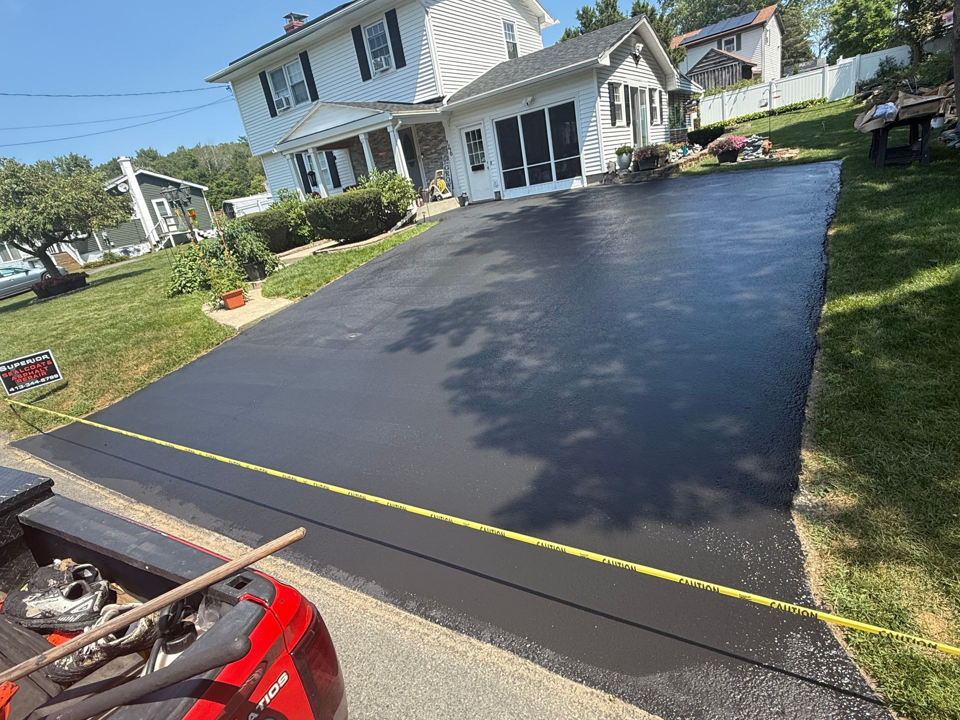 A freshly sealed, smooth black driveway in front of a white two-story house, viewed from the back of a red truck.