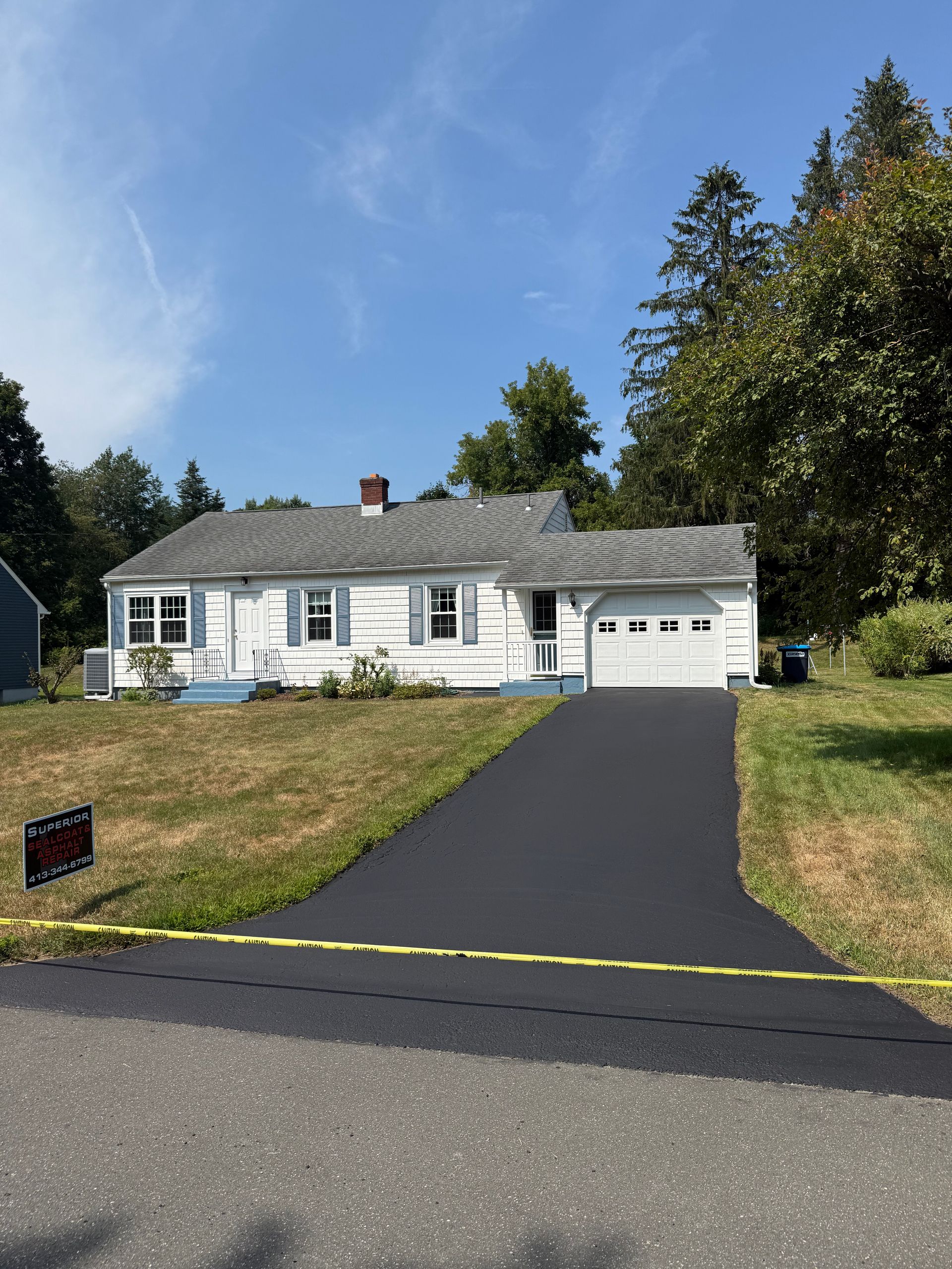 A single-story white house with a gray roof and an attached garage, featuring a newly paved dark asphalt driveway.