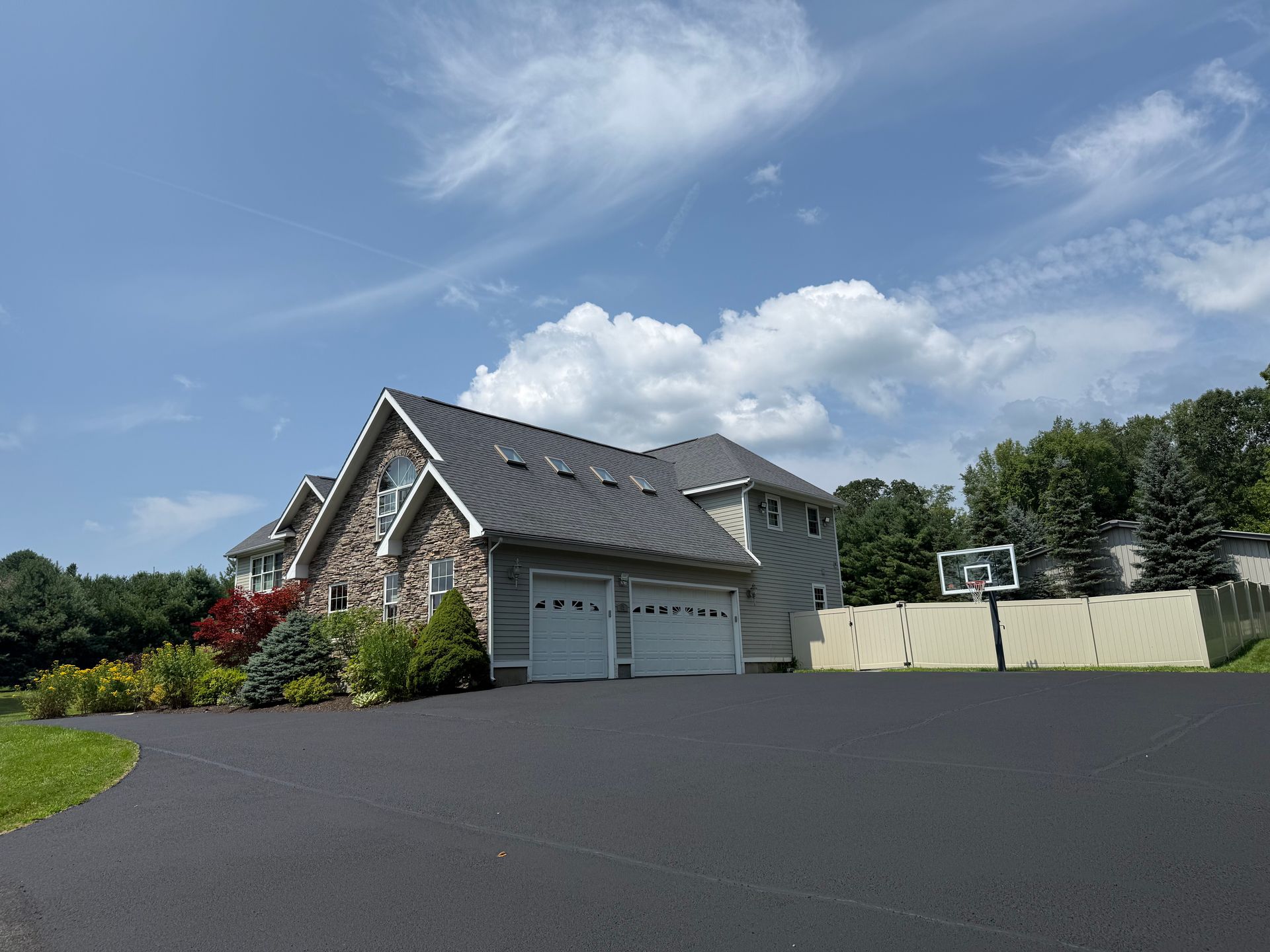 A multi-story stone and siding house with a metal roof and a large asphalt driveway, featuring a backyard basketball hoop.