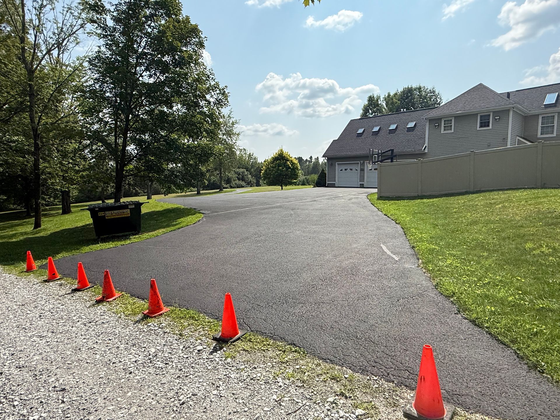 A line of orange traffic cones separates a gravel area from a newly paved asphalt driveway leading to a suburban house.