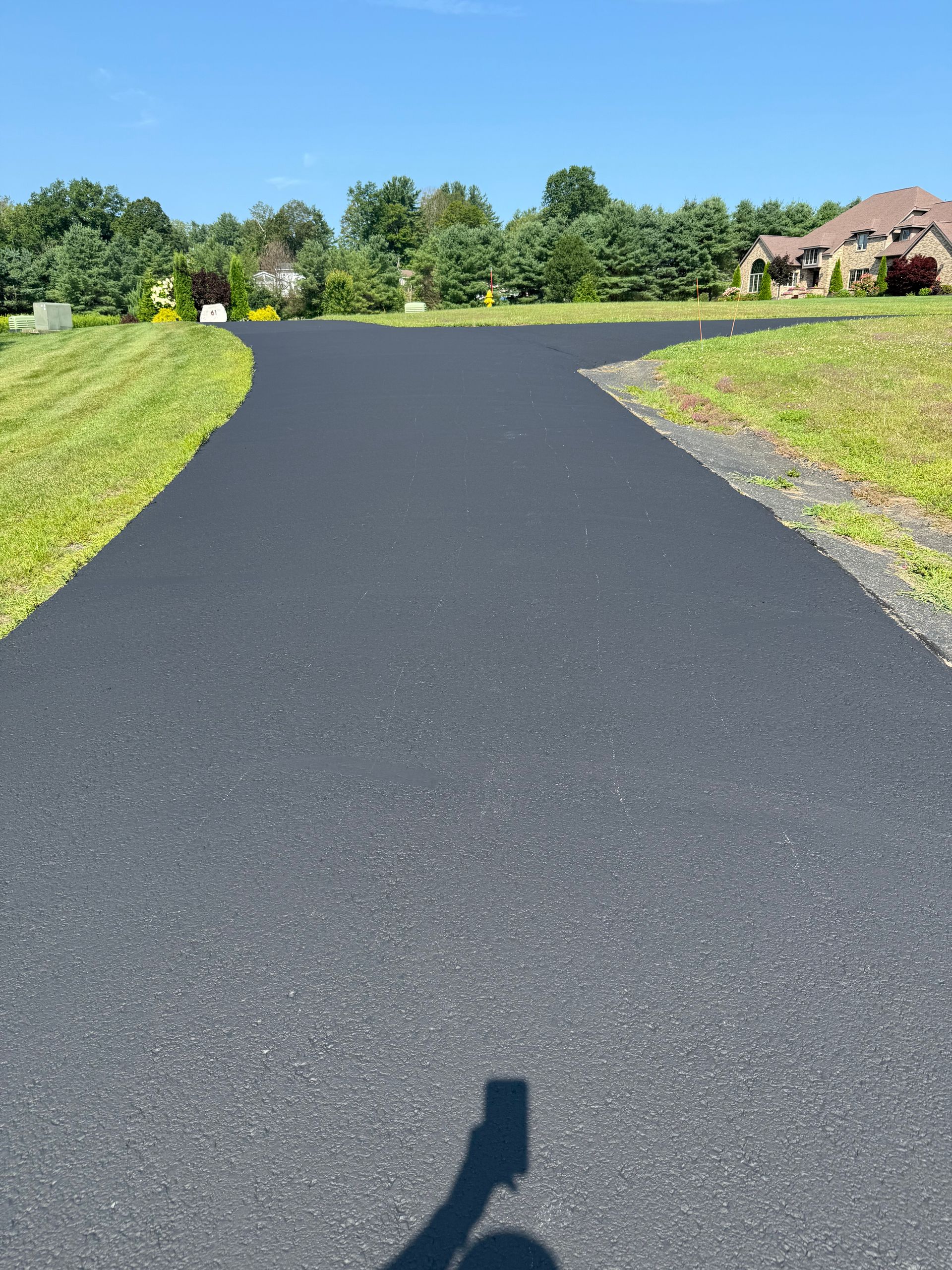 A paved driveway forks in two directions through a grassy yard toward a house in the distance under a clear blue sky.