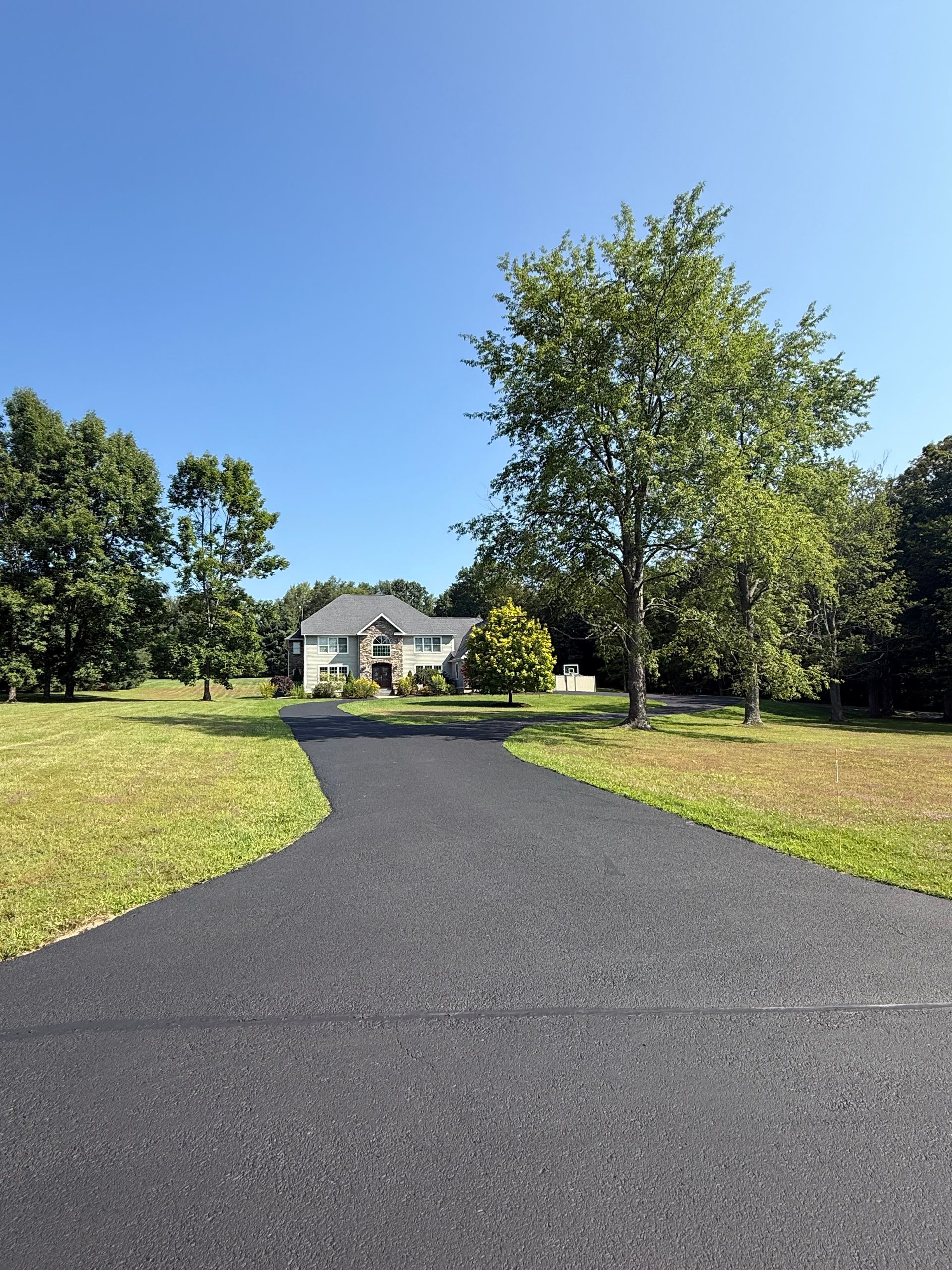 A long, freshly paved black asphalt driveway leads toward a house in the distance surrounded by green trees and grass.