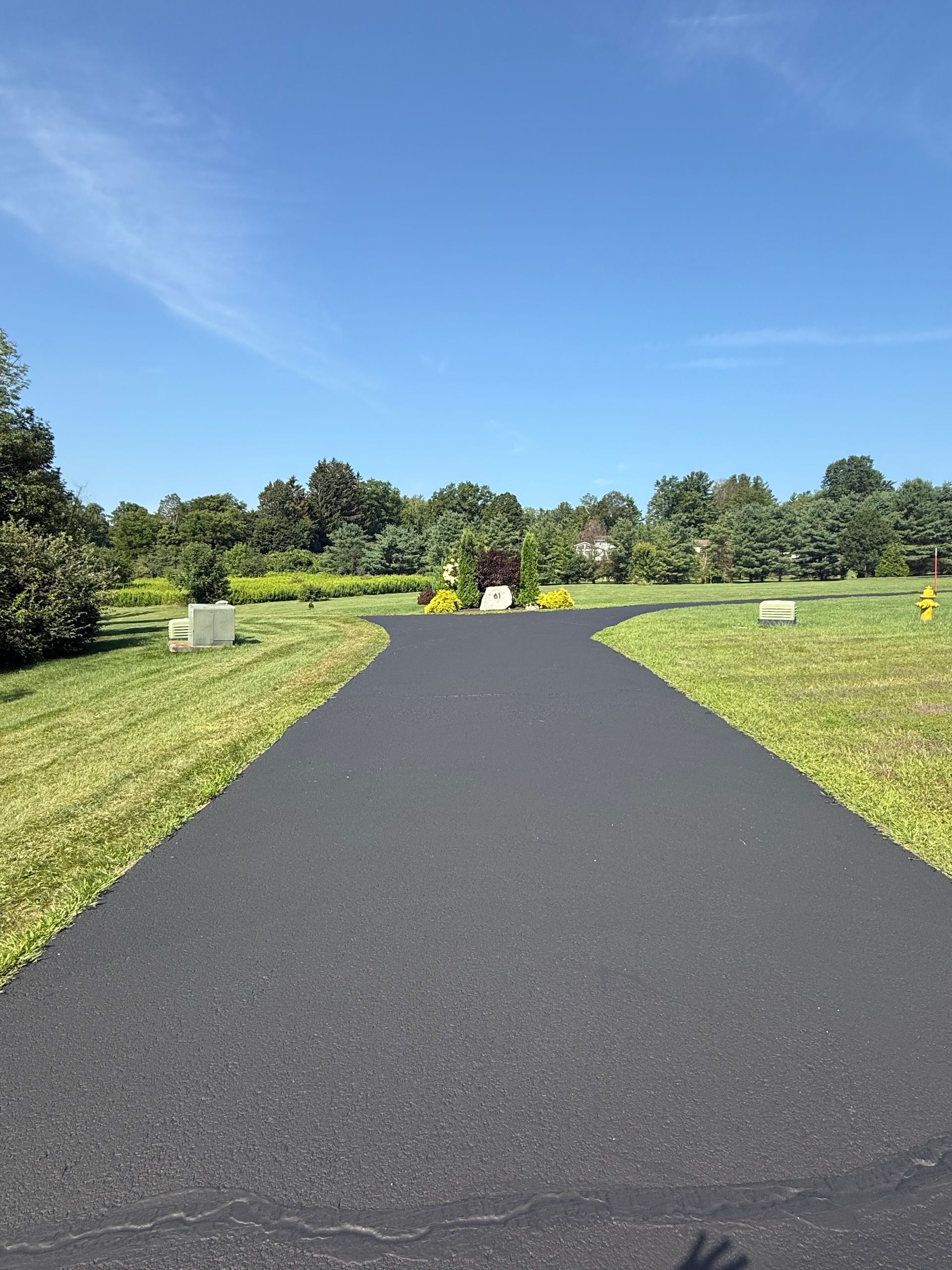 A freshly paved asphalt path leads toward a sunlit memorial garden under a clear blue sky.