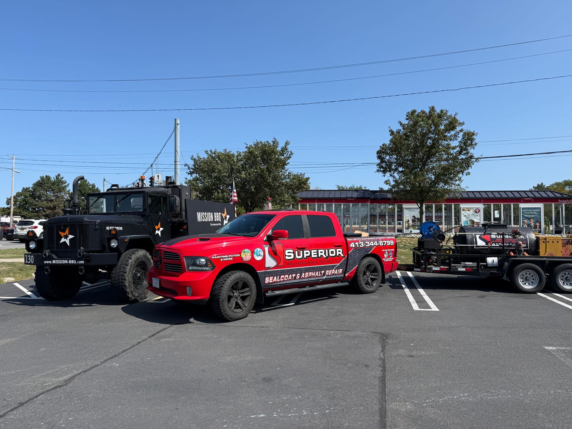 A black military-style truck parked next to a red pickup truck pulling a trailer in a sunny paved parking lot.