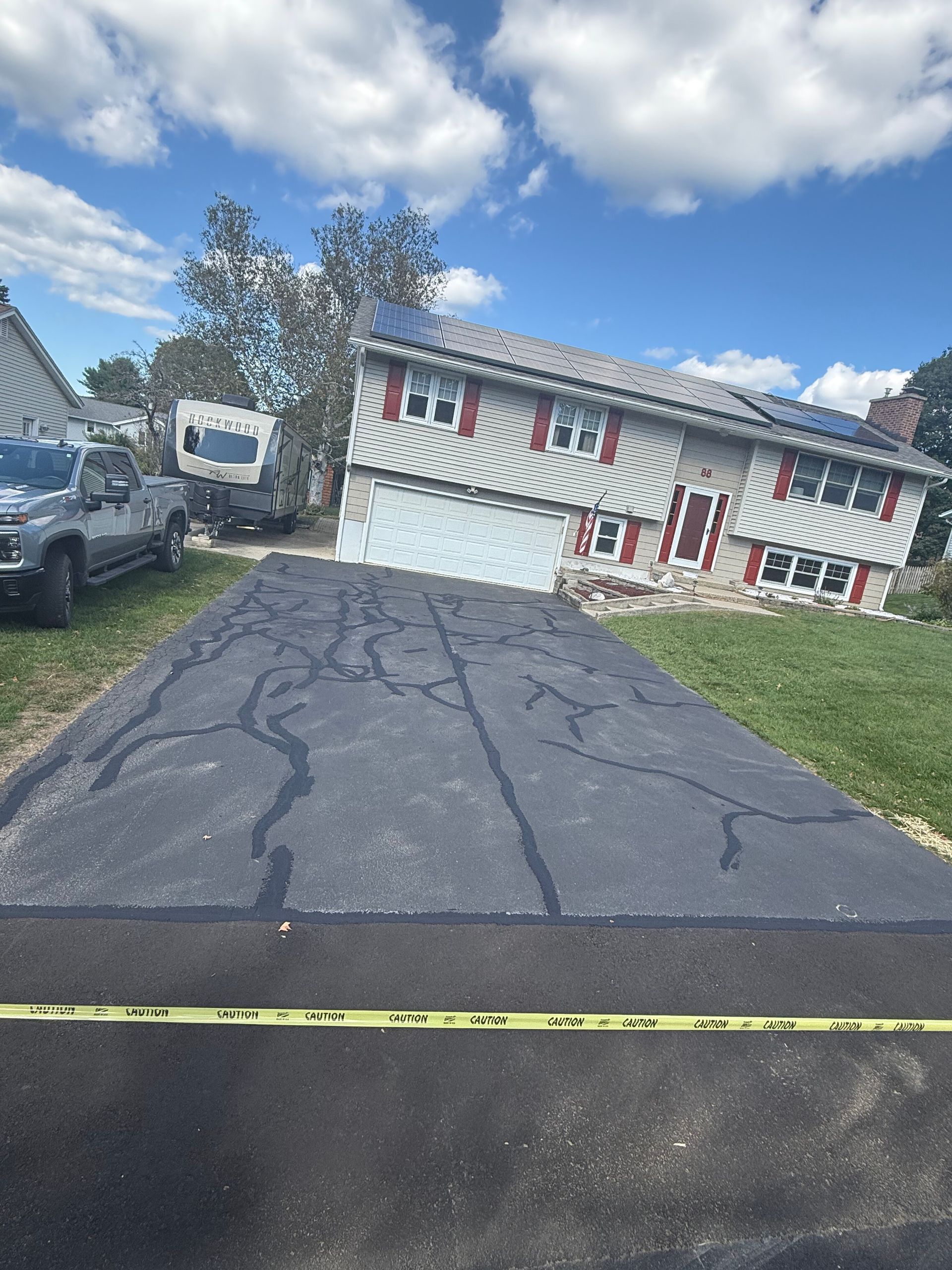 A house with red shutters sits behind a paved driveway marked with caution tape and filled with dark crack-filler lines.
